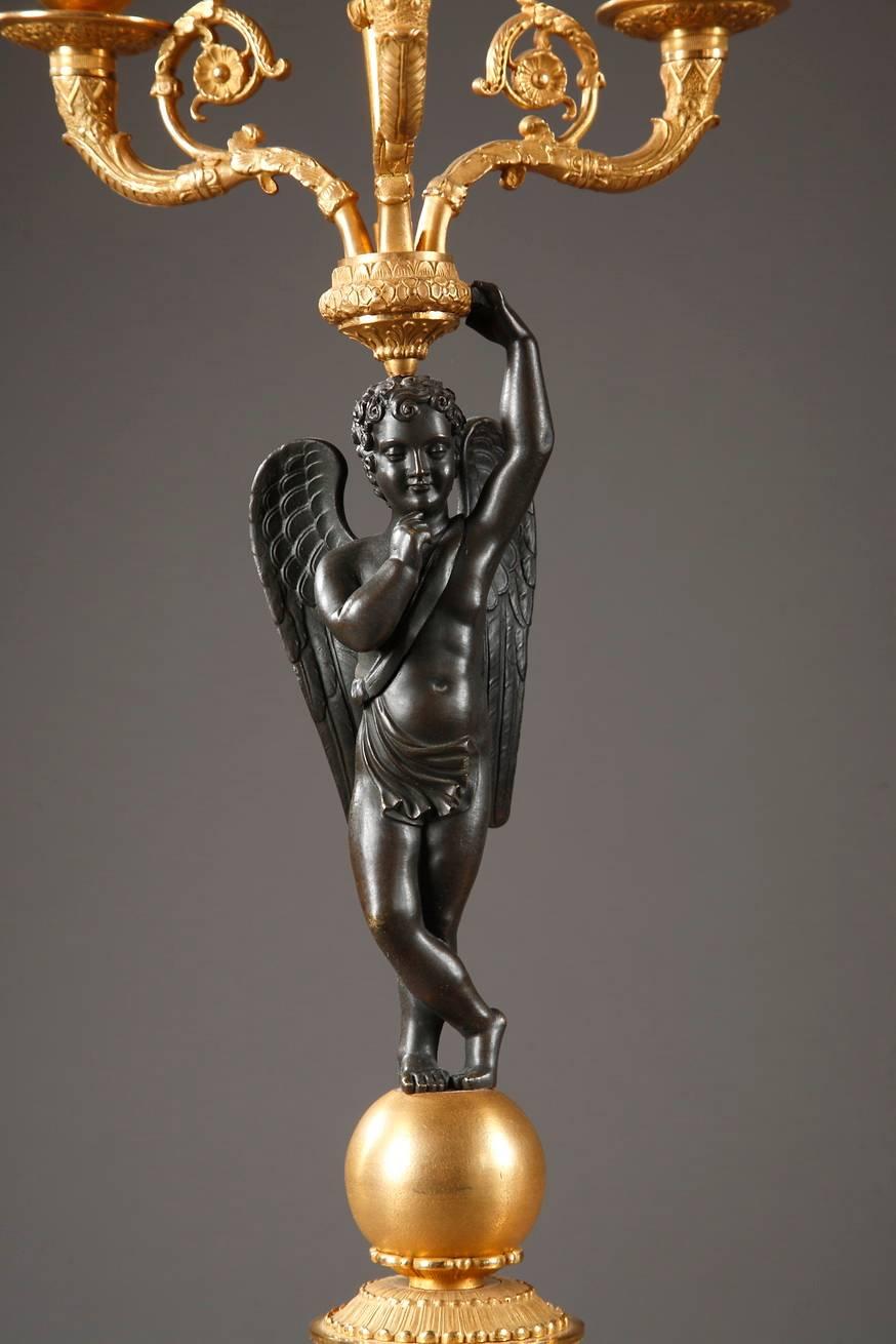 Impresionante pareja de candelabros de bronce dorado y patinado, con un cupido alado. He está de pie sobre una esfera y sostiene una antorcha, que está rodeada por tres ramas de vela curvadas. Las ramas están decoradas con flores y follaje en