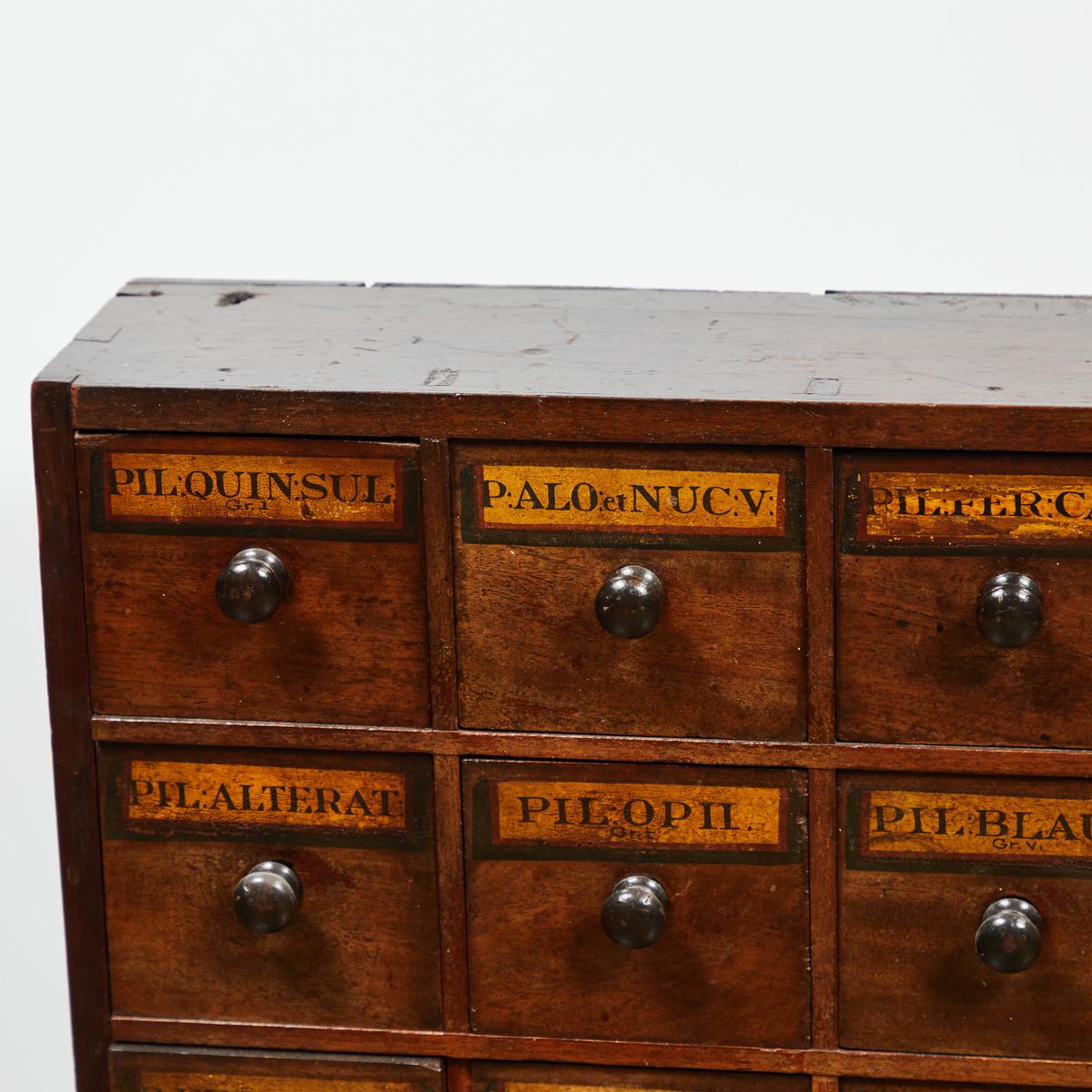 Early 19th century English apothecary wall chest with hand written labels, to hang or place on stand. The narrow rectangular body holds 28 square drawers with wooden knob handles.