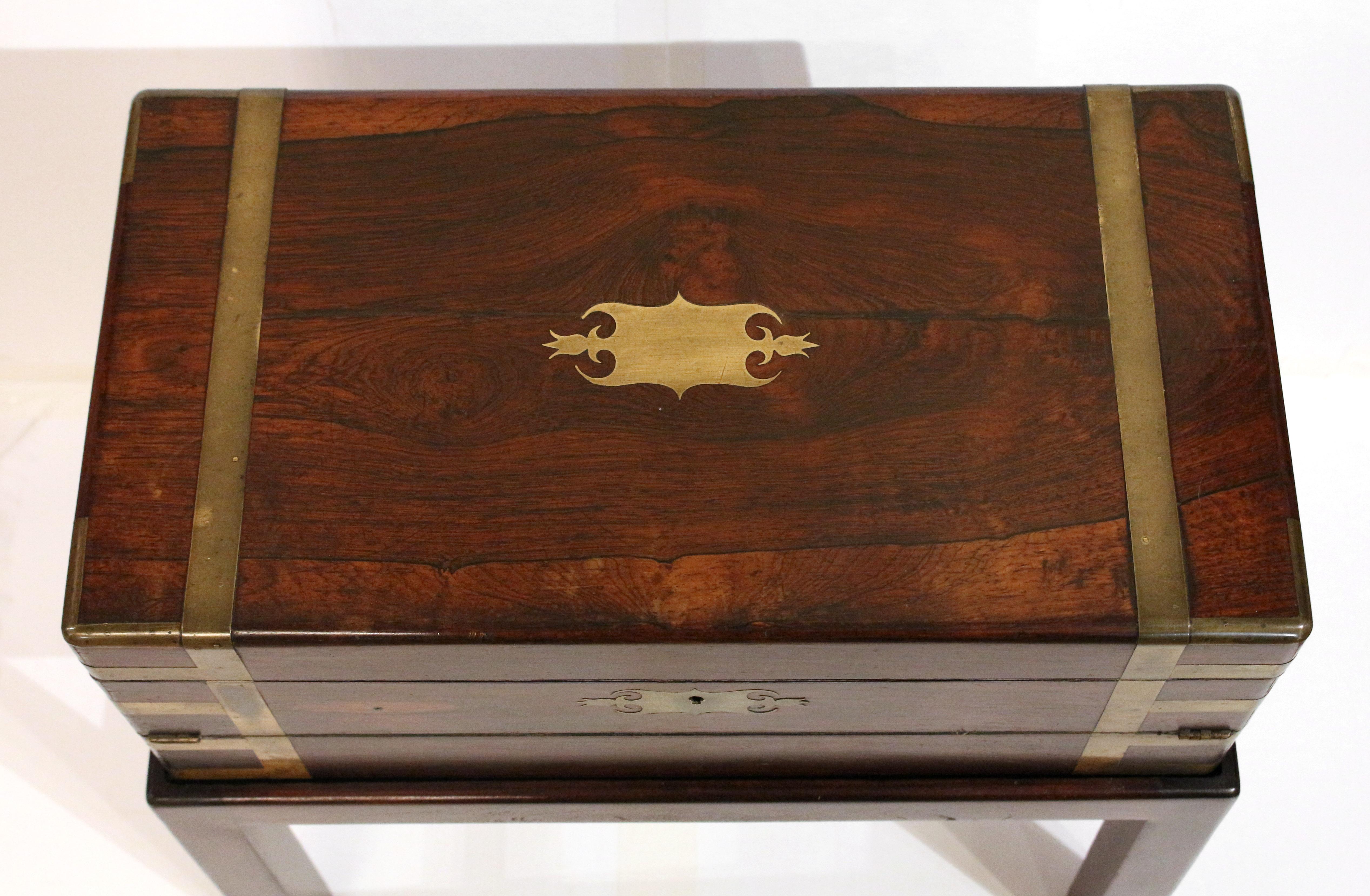 Early 19th century double fold campaign lap desk box on stand side table, English. A rarely found form. George III to Regency period. Rosewood. Gilt tooled black leather interior surfaces. Two inkwells. Extensive brass strapping, cartouche