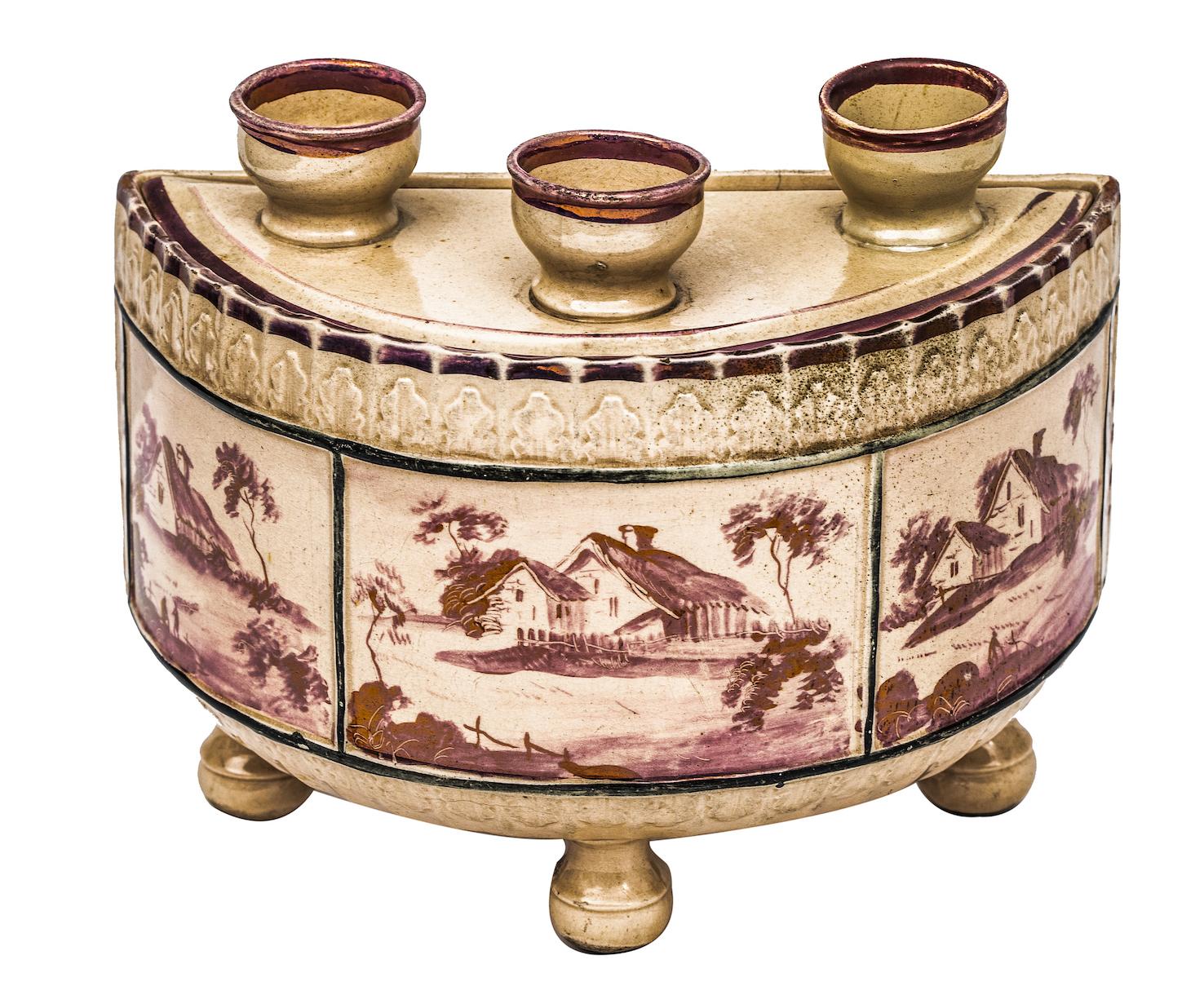 This demilune flower pot or bough pot for forcing springtime bulbs, is adorned with three landscape panels. Each panel, painted in pink lustre, depicts the same cottage in a pastoral setting from three different vantage points. Moulded into the top