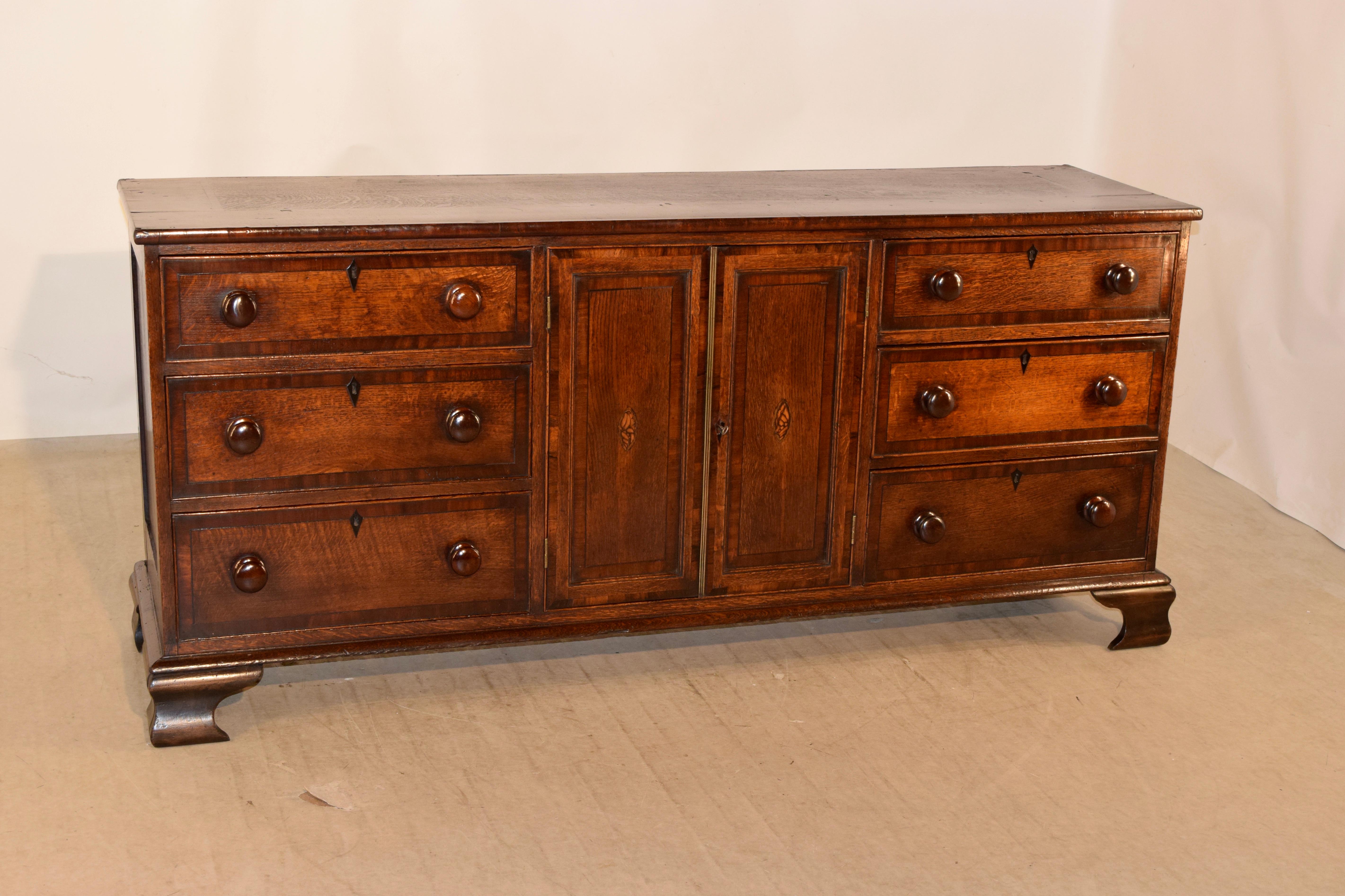 Early 19th century English oak dresser base with wide mahogany band around the top following down to paneled sides and a pair of central paneled doors with shell inlay, which open to reveal shelving. The doors are flanked on either side by three