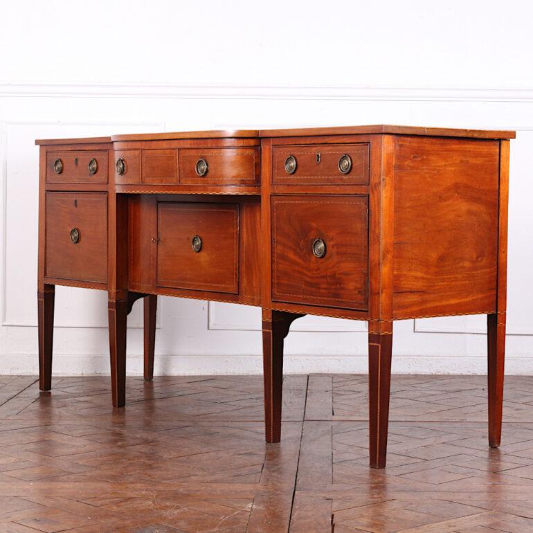 An early-19th century English buffet in mahogany, fitted with a variety of drawers and cabinets including smaller drawers under the top and a large 'cellaret' drawer to the lower left. The piece features decorative inlaid stringing and banding to