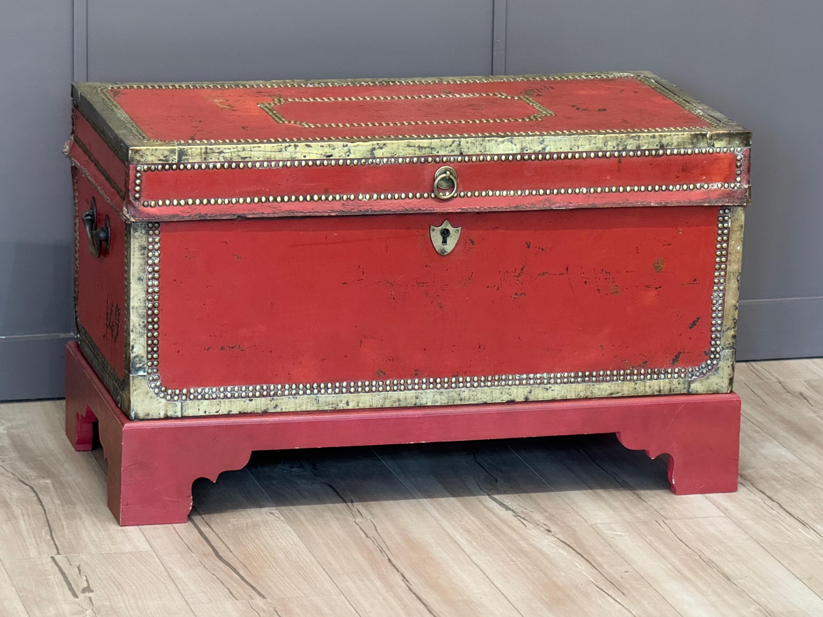 An English traveling trunk clad in vivid red leather and trimmed with brass tacks and banding. Raised on a custom red-painted base with shaped bracket feet, the trunk features side carrying handles and a ring-pull latch. The interior retains its