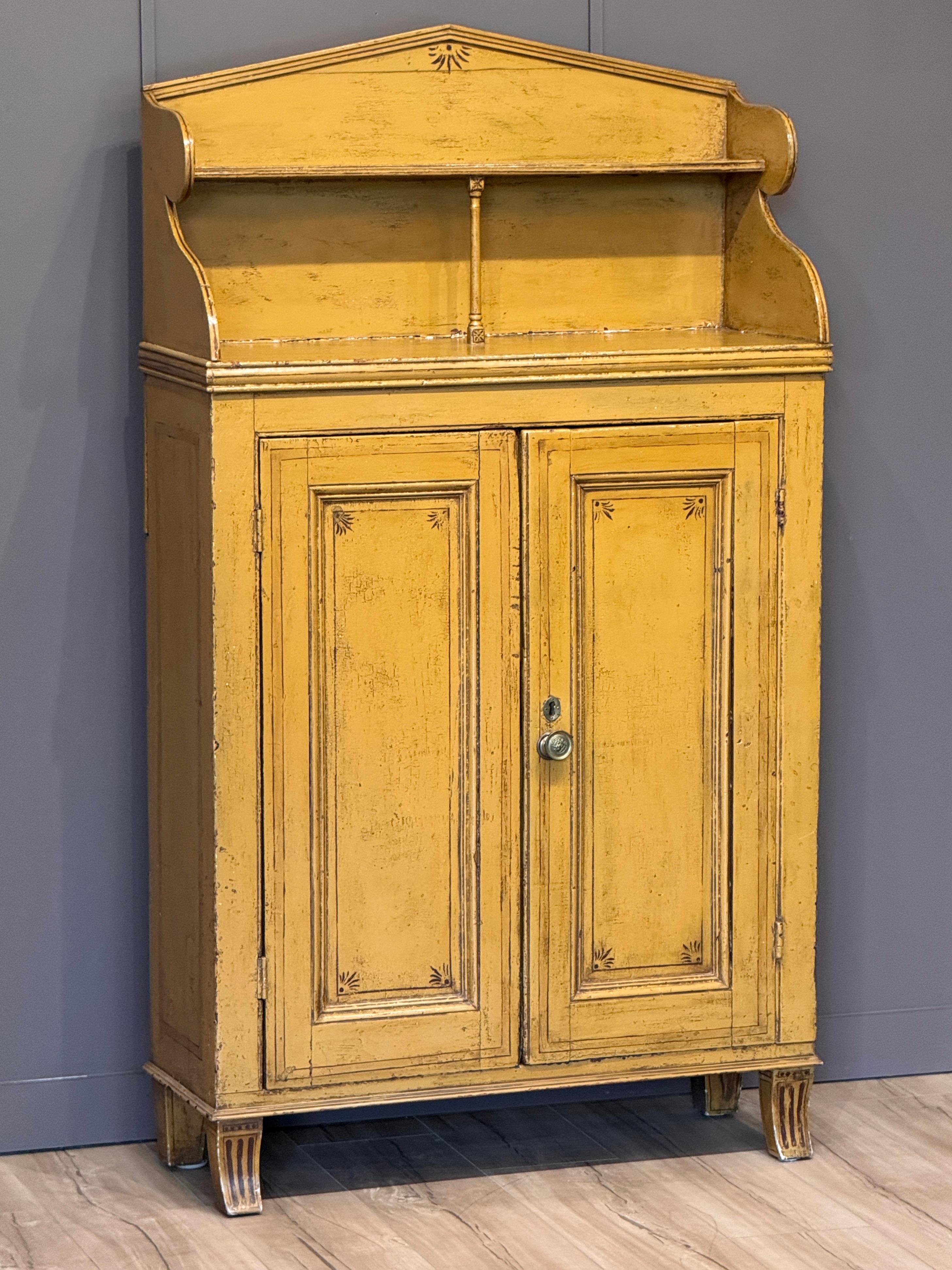 Un mobile chiffonier inglese dell'inizio del XIX secolo con decorazioni originali in giallo senape. Il piano architettonico presenta lati sagomati e un ripiano centrale sostenuto da una colonna tornita. La cassa è dotata di ante a pannelli e poggia