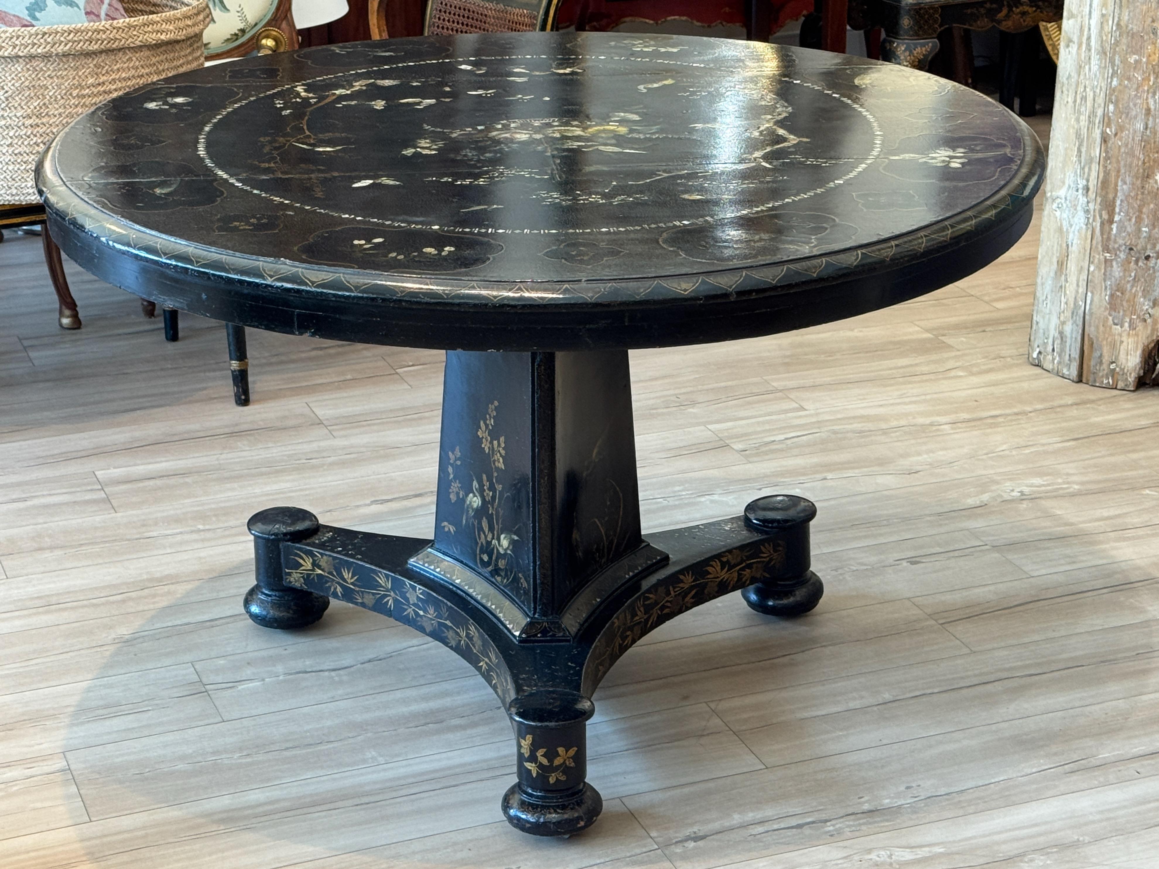 An early 19th century English Regency tilt-top center table in black lacquer, decorated with polychrome chinoiserie-style motifs and mother-of-pearl inlay. The circular top rests on a tapering triangular pedestal, raised on a tripartite base with