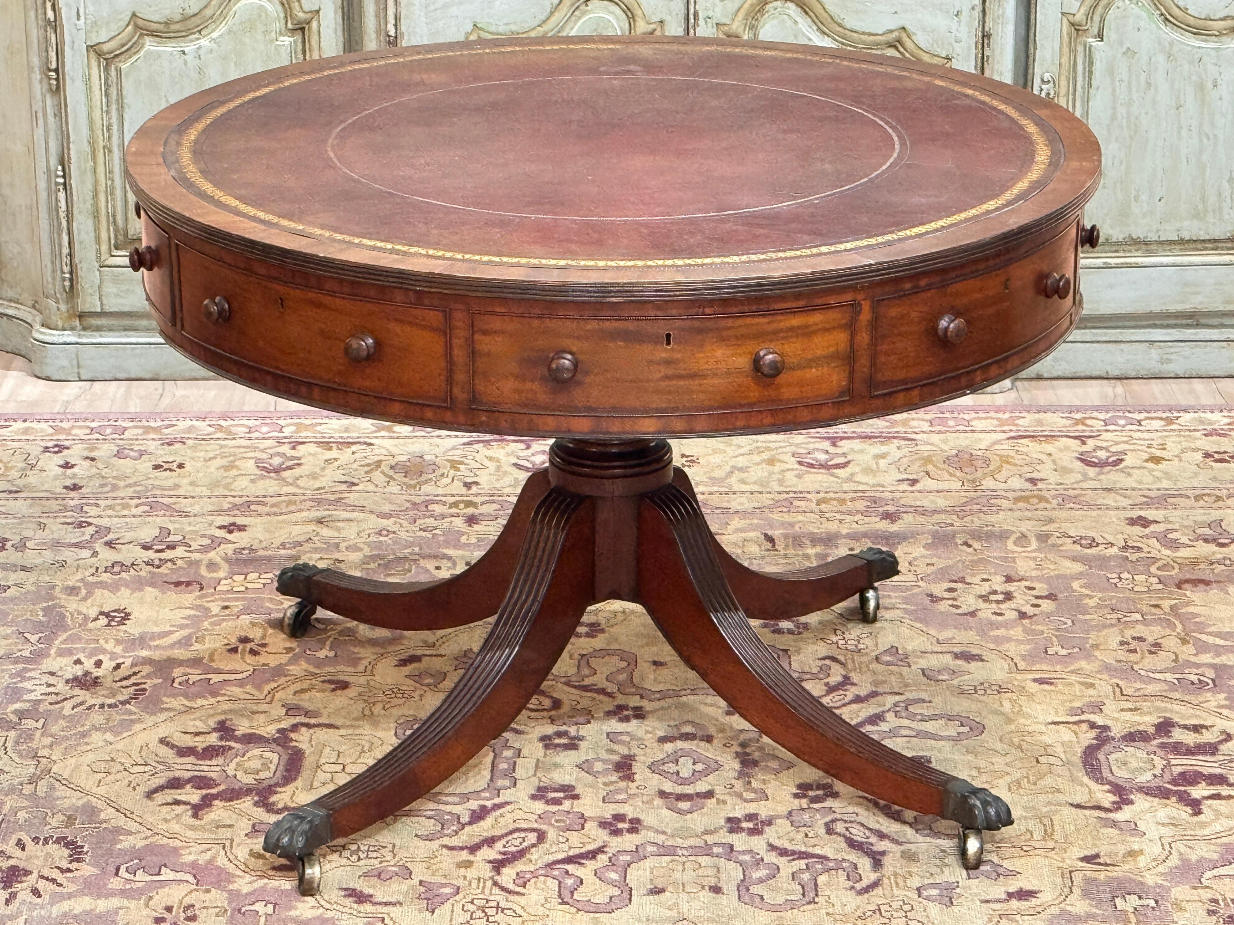 An early 19th century English Regency drum table in mahogany with inset tooled leather top and a band of real and faux drawers around the apron. Raised on a turned pedestal with reeded, saber-form legs ending in brass paw feet and casters. Dovetail