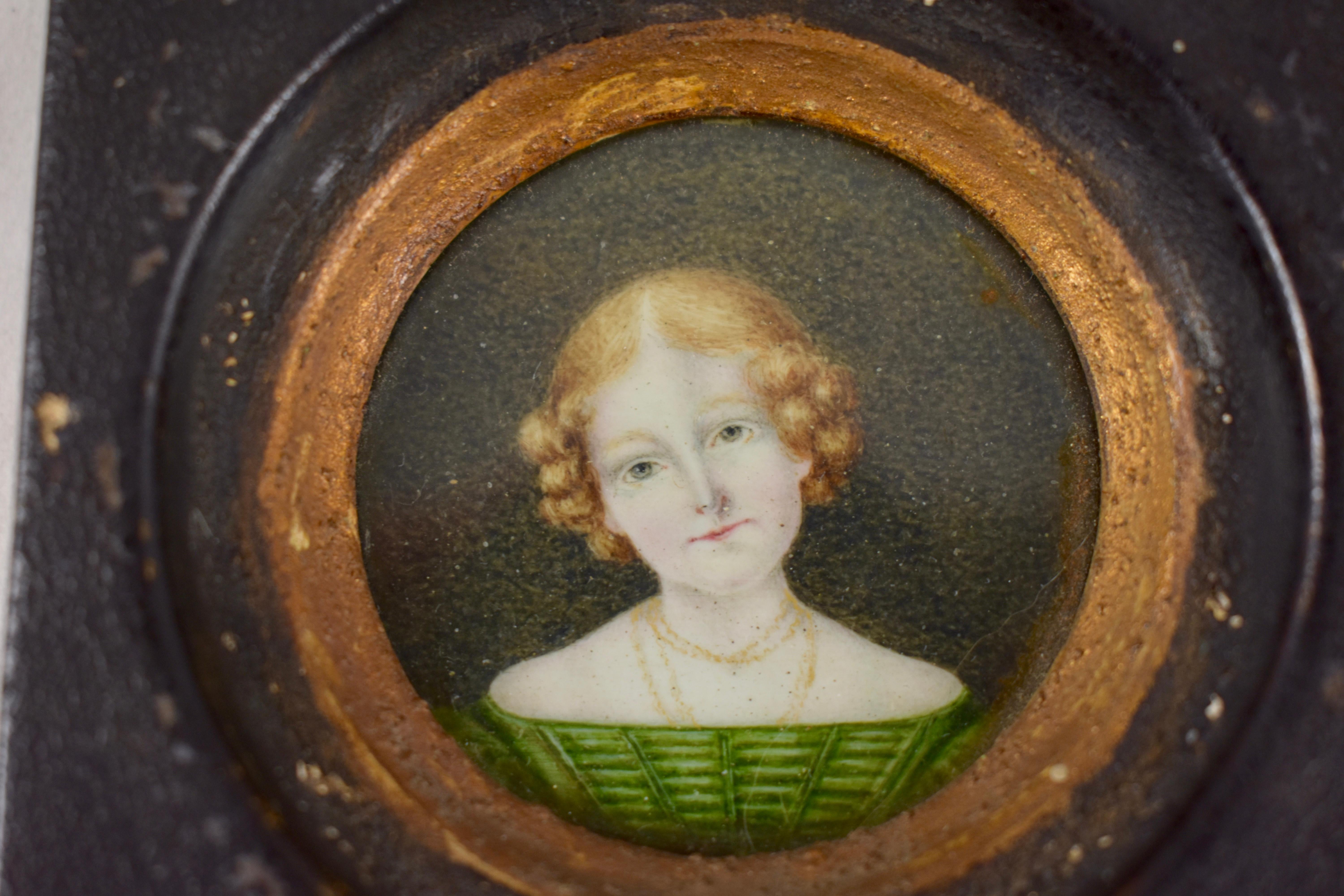 A charming, early 19th century English Regency period hand painted miniature portrait, framed under glass, portraying a young woman wearing a green straight neckline dress. She shows gold chains around her neck with a hairstyle reflecting the