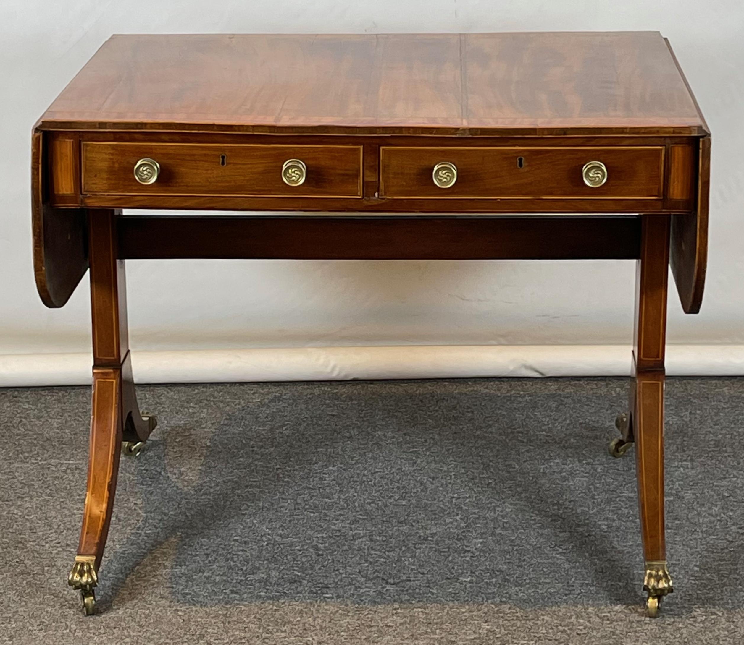 A small and elegant sun-faded mahogany Regency sofa table accented with satinwood and ebony resting on down swept saber legs terminating in brass hairy paw casters.