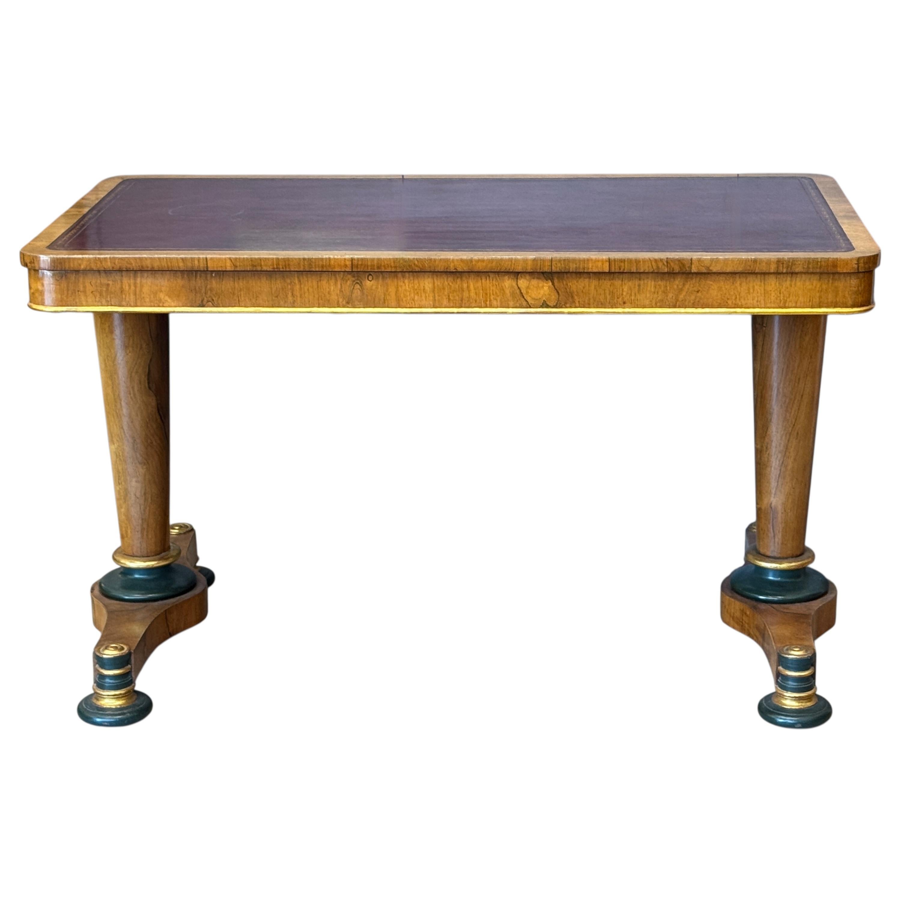 An early 19th century English library table in rosewood, featuring an inset red tooled leather top above a rectangular apron. The twin pedestal base is supported by turned columns resting on shaped legs with ebonized and gilt accents, terminating in