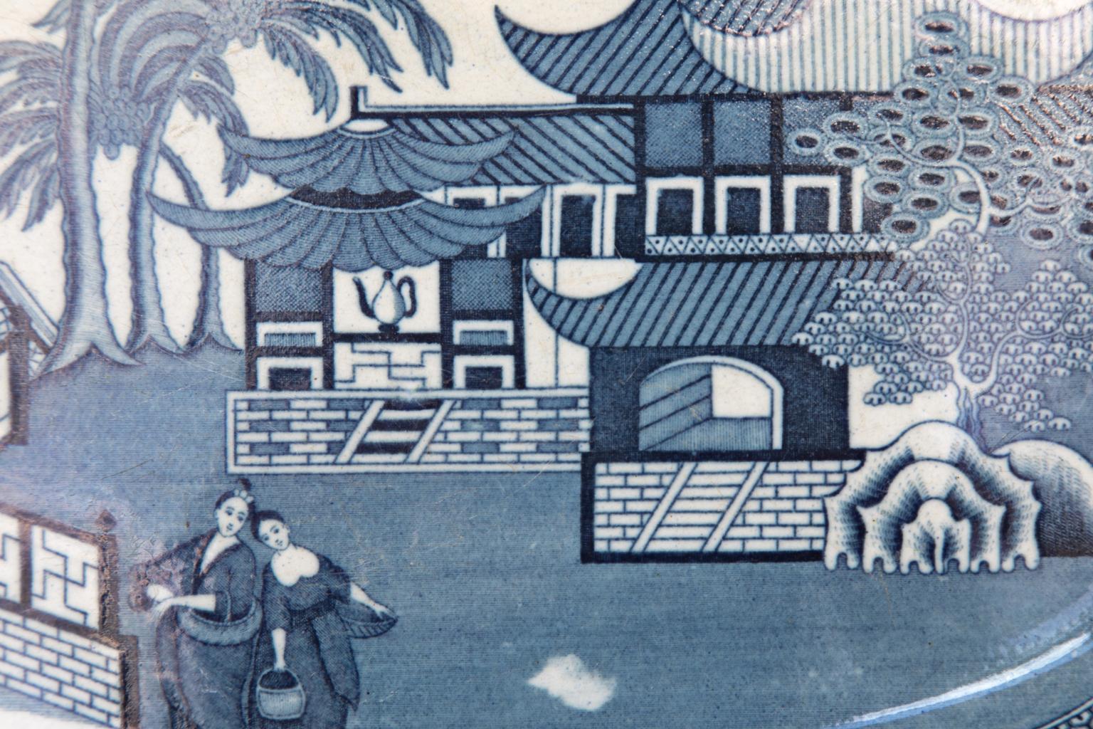 Early 19th century English staffordshire blue and white platter, with a Chinese landscape featuring houses, female figures, fences, bridges, and coconut palm trees. The border is has a floral design.