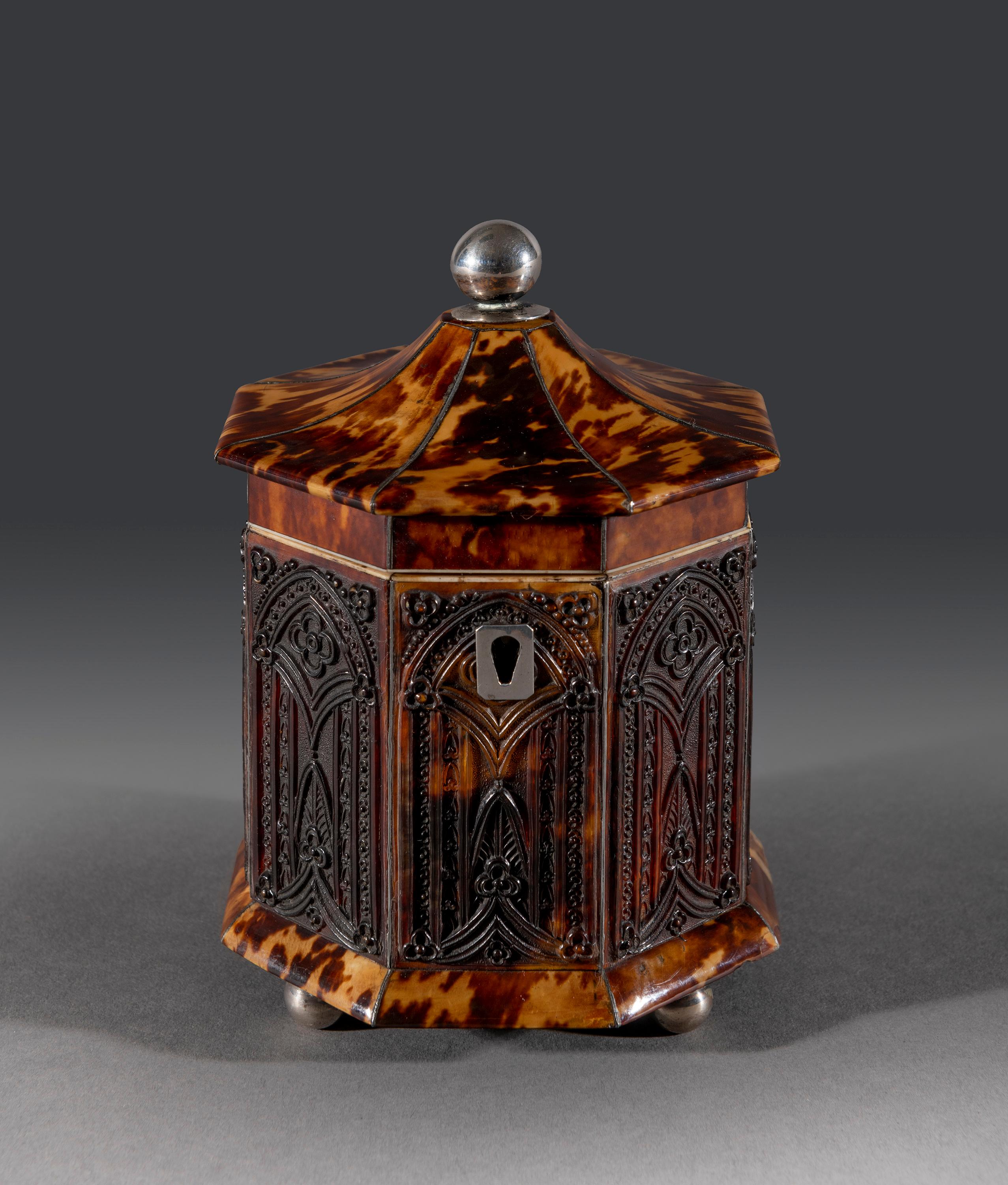 The small octagonal tortoiseshell caddy has a silver ball finial and escutcheon with octagonal pressed Gothic arches to each side separated with silver stringing. The caddy sits on four silver ball feet. The top opens to reveal a single bore handle