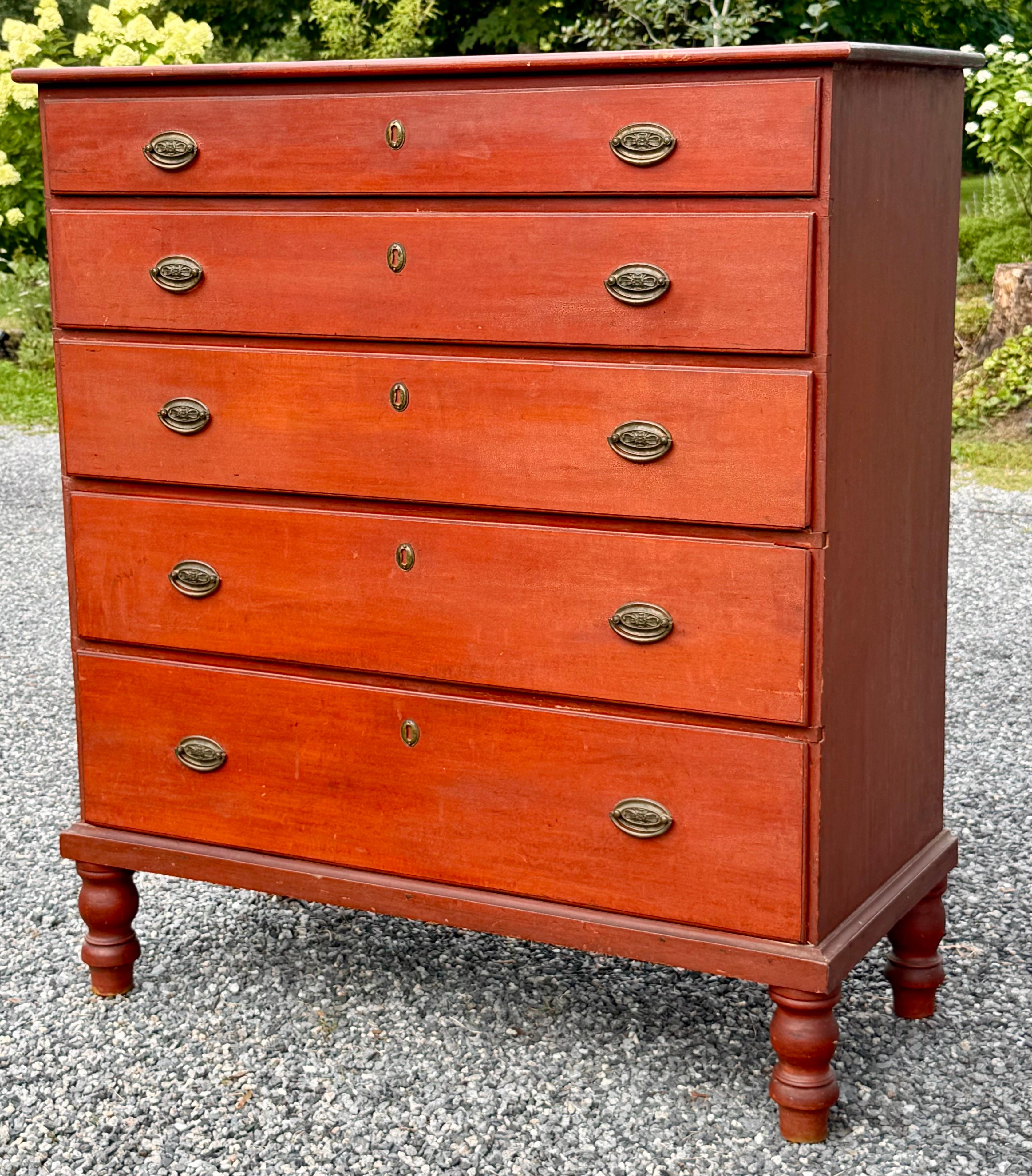 An early 19th century New England Federal period tall chest in old red paint featuring five graduated dovetailed drawers and retaining original brasses.  Molded base with heavy turned legs measuring 45”H x 40”W x 18.5”D.  Presents very well overall