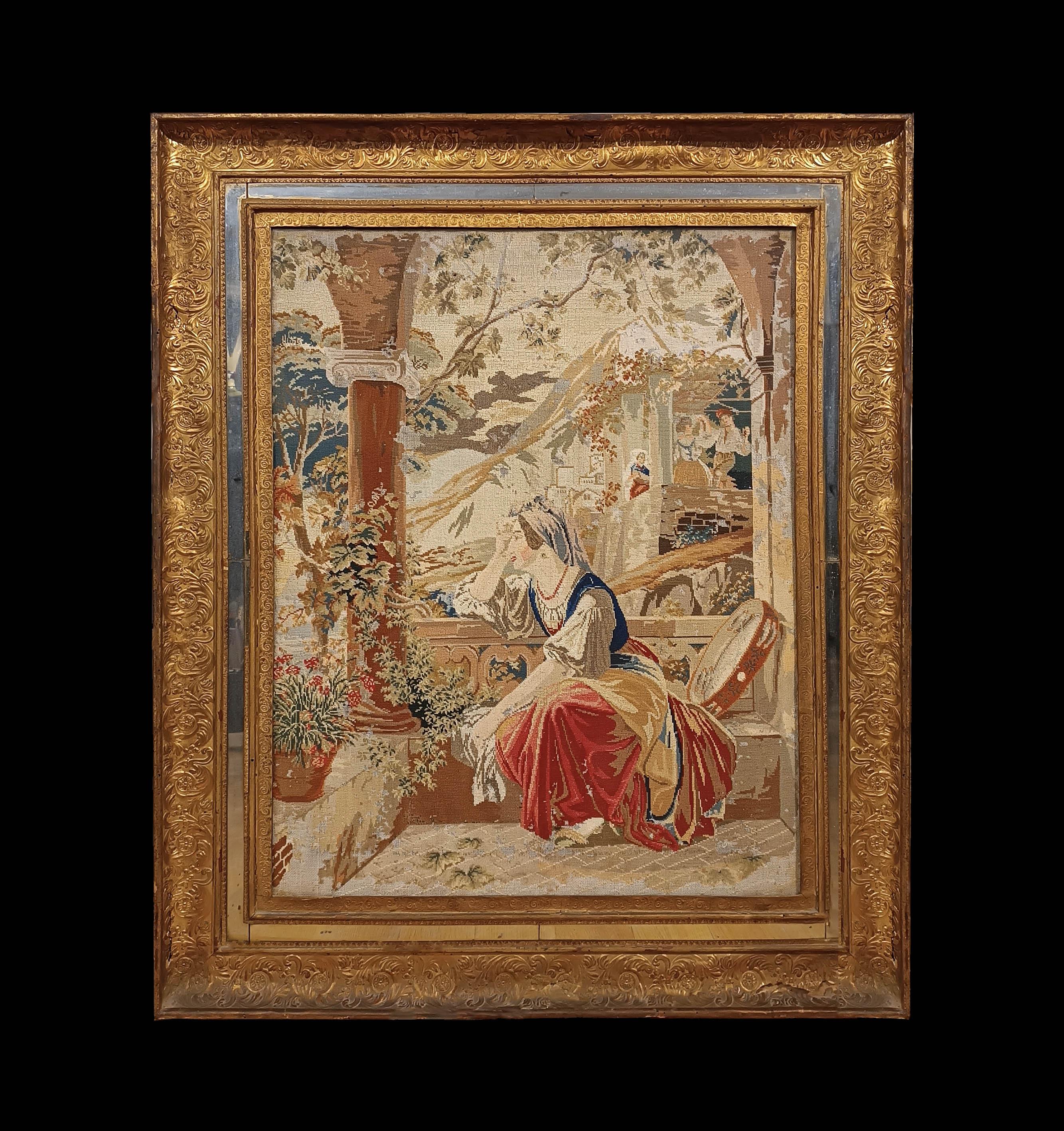Beautiful small stitch embroidered fabric framed in a carved and gilded frame, with a small mirrored part. The embroidery depicts a romantic scene, with a woman sitting on the balcony of a terrace with columns and plants. The woman wears brightly