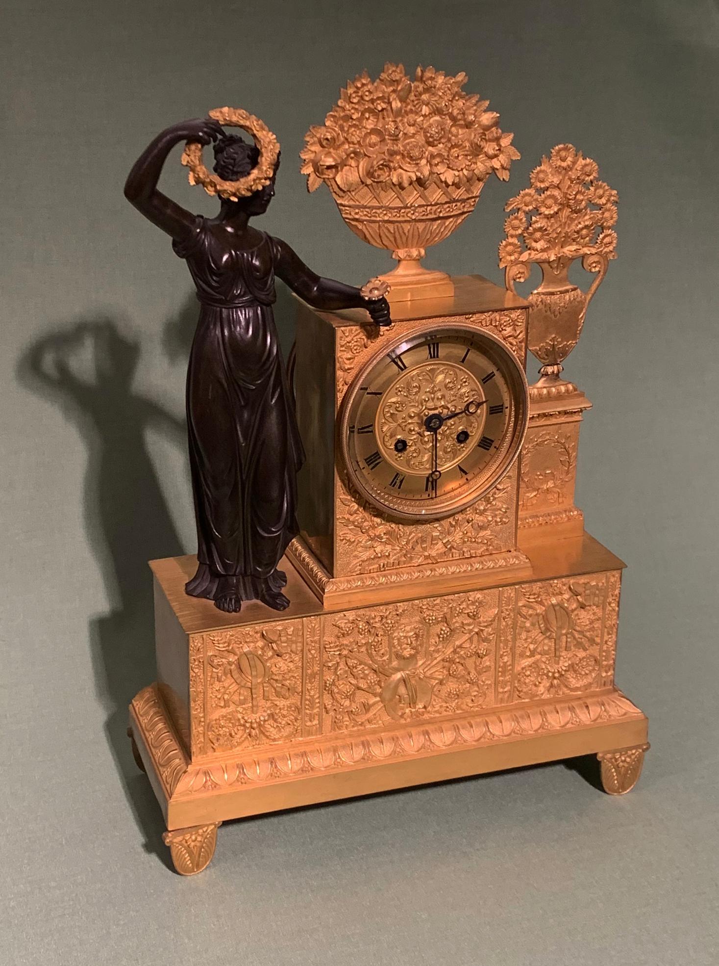 An early 19th century French eight day silk-suspension, striking clock, contained in ormolu case mounted with vases and urns of flowers with well-cast bronze figure of a muse holding a wreath of flowers, all supported on plinth engraved with central