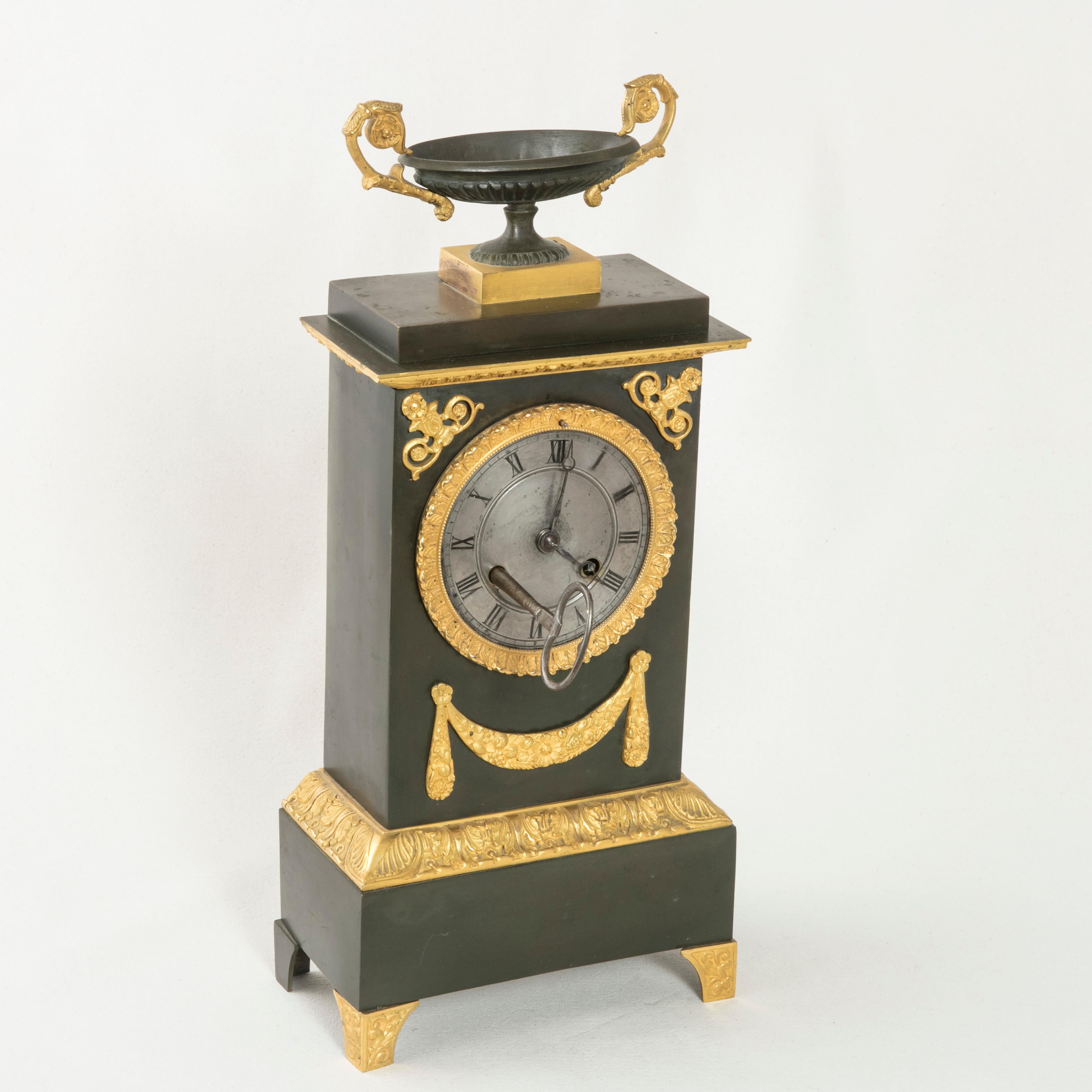 This early 19th century French Charles X period iron mantel (fireplace) clock features gilt bronze detailing of scrolling and a swag below the face. An iron urn with gilt bronze handles crowns the top and stands on a bronze plinth. Gilt bronze trim