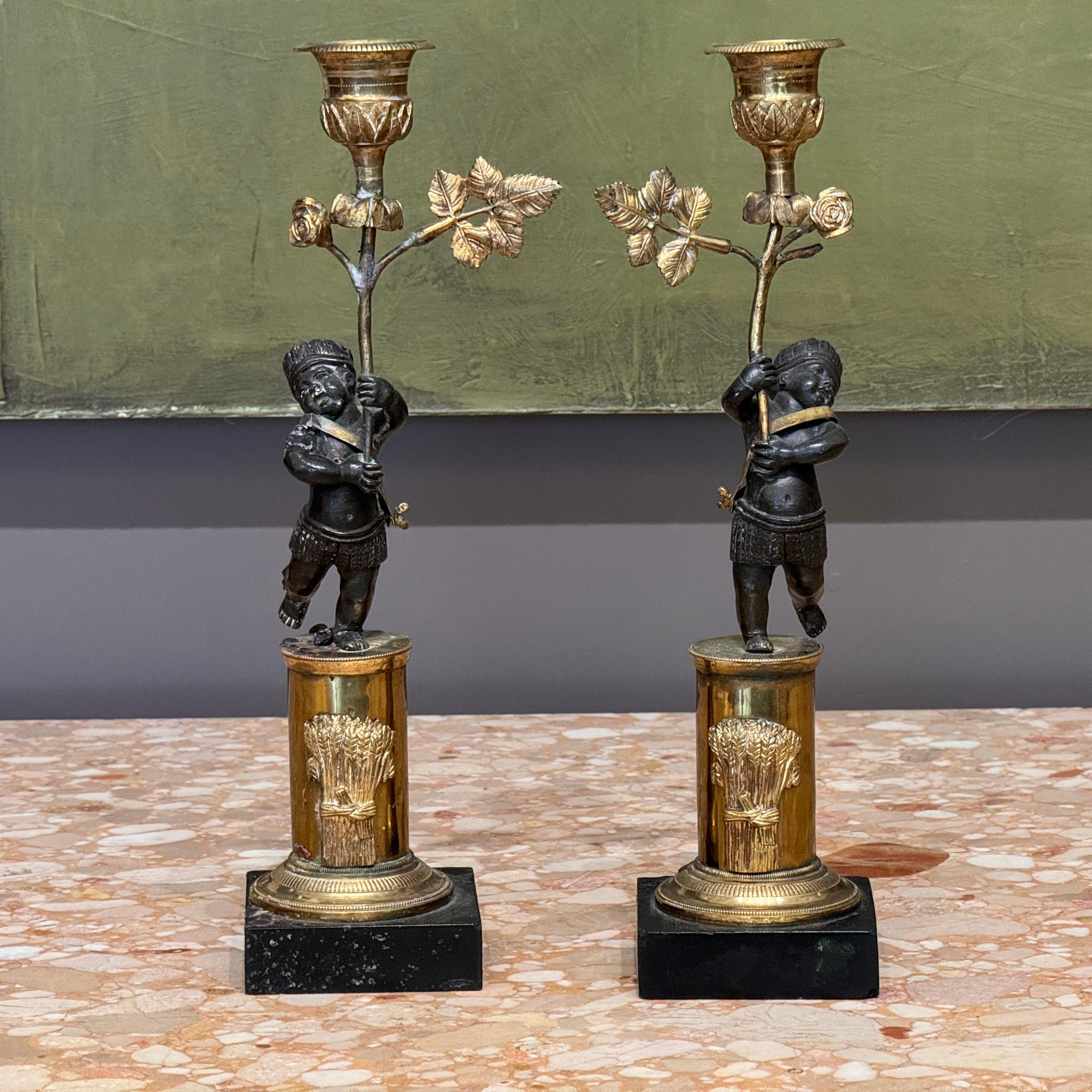 Pair of rare early 19th century French Empire figural bronze candlesticks featuring patinated putti figures dressed in stylized costume, each supporting a foliate ormolu candle branch. The cylindrical gilt bronze columns are mounted with sheaves of