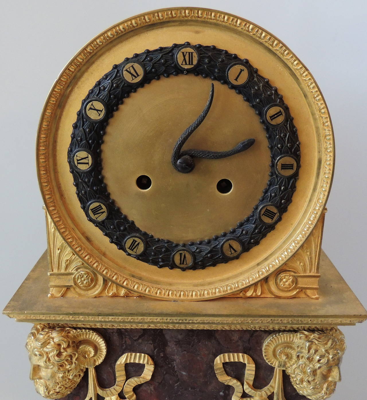 This early 19th century Empire style figural clock with a snake as the hands on the clock is set on wonderful rouge marble and a pedestal base. The four corners are marked with four wonderfully detailed doré bronze figure heads, The front is
