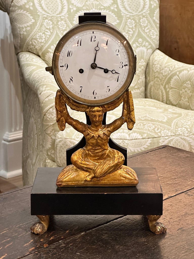 An early 19th C. French carved gilt wood mantle clock, depicting a fancifully dressed, cross-legged figure, supporting the clock, resting on an ebonized wood base with carved, gilded feet.