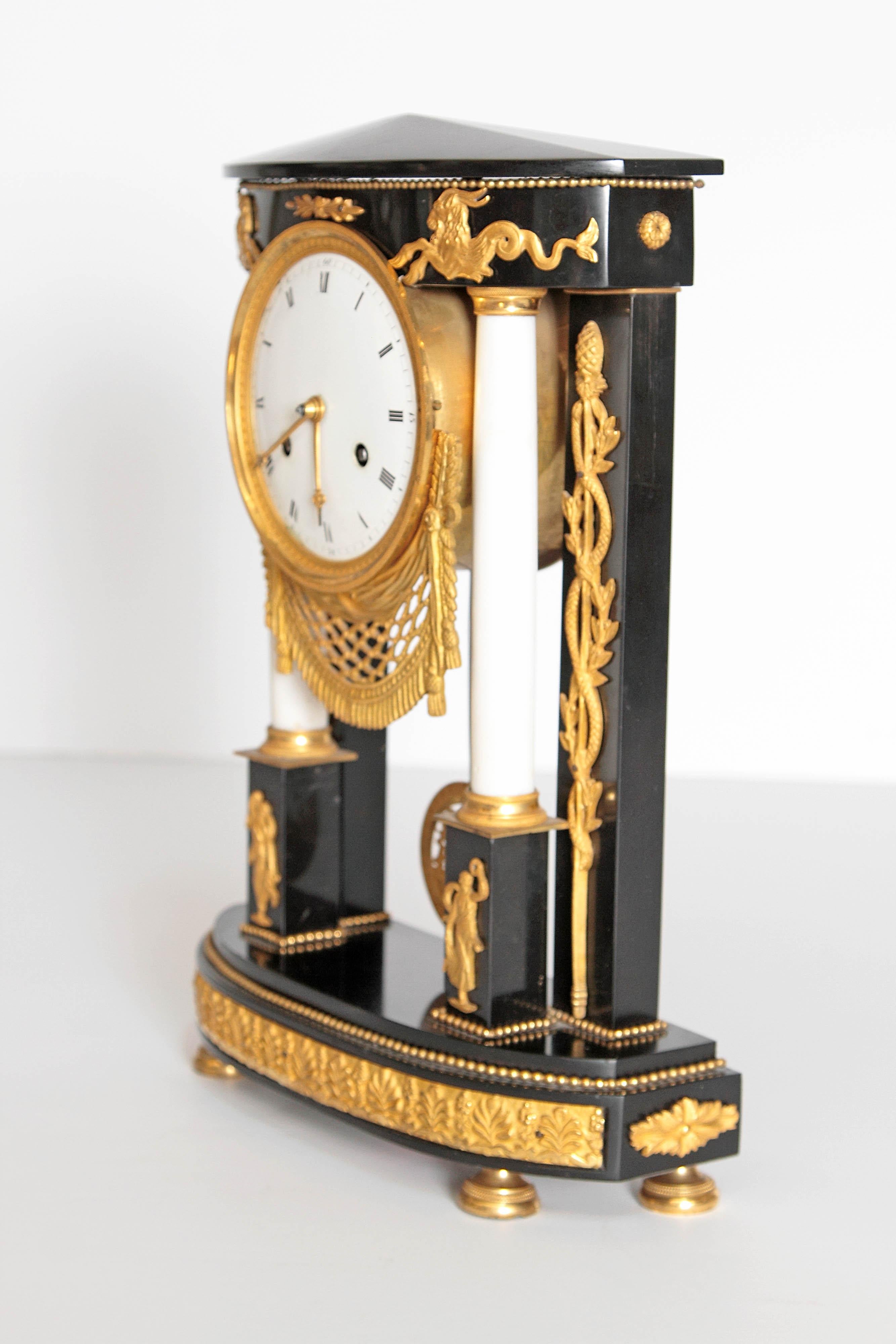 A French black marble neoclassic clock. Square columns at back with round white columns on pedestals framing round clock face. Delicate gilt bronze mounts embellish the whole. Serpentine twists at the sides, small ram figures at top of columns and