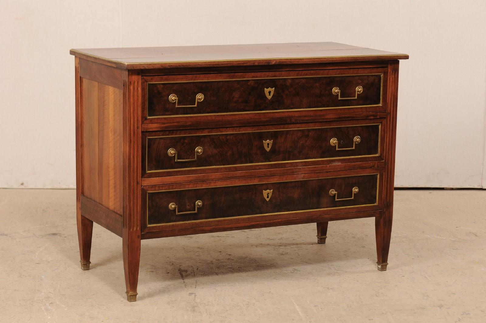 A sophisticated French neoclassical style chest of three drawers, from the early 19th century, with contrasting woods and brass trim. This antique chest from France features a rectangular-shaped top with brass trim edging, atop a neoclassical case