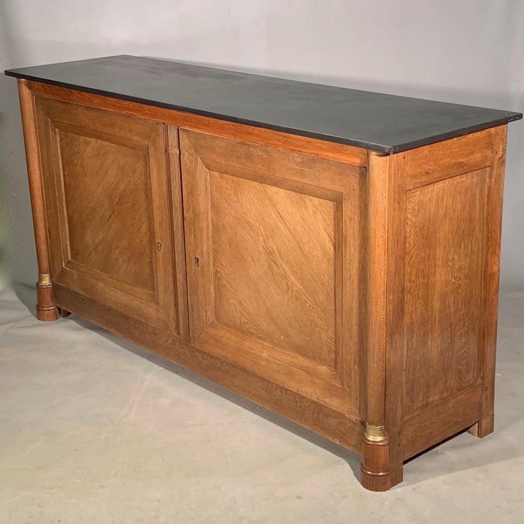 Unusual early 19th century French Empire period solid oak sideboard with a slate top.
Lovely simple style with the panelled doors and sides, flanked with the side columns and finished with brass collars top and bottom.
The whole sideboard is made