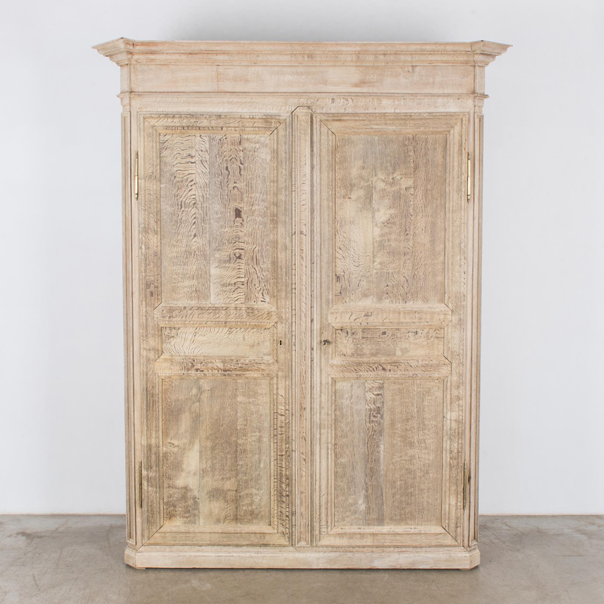 A bleached oak armoire from France, circa 1800. Two paneled doors open into a pair of interior shelves. The angled casing possesses a stately, neoclassical sensibility. Molded cornices give a sense of grandeur. The doors close with an original