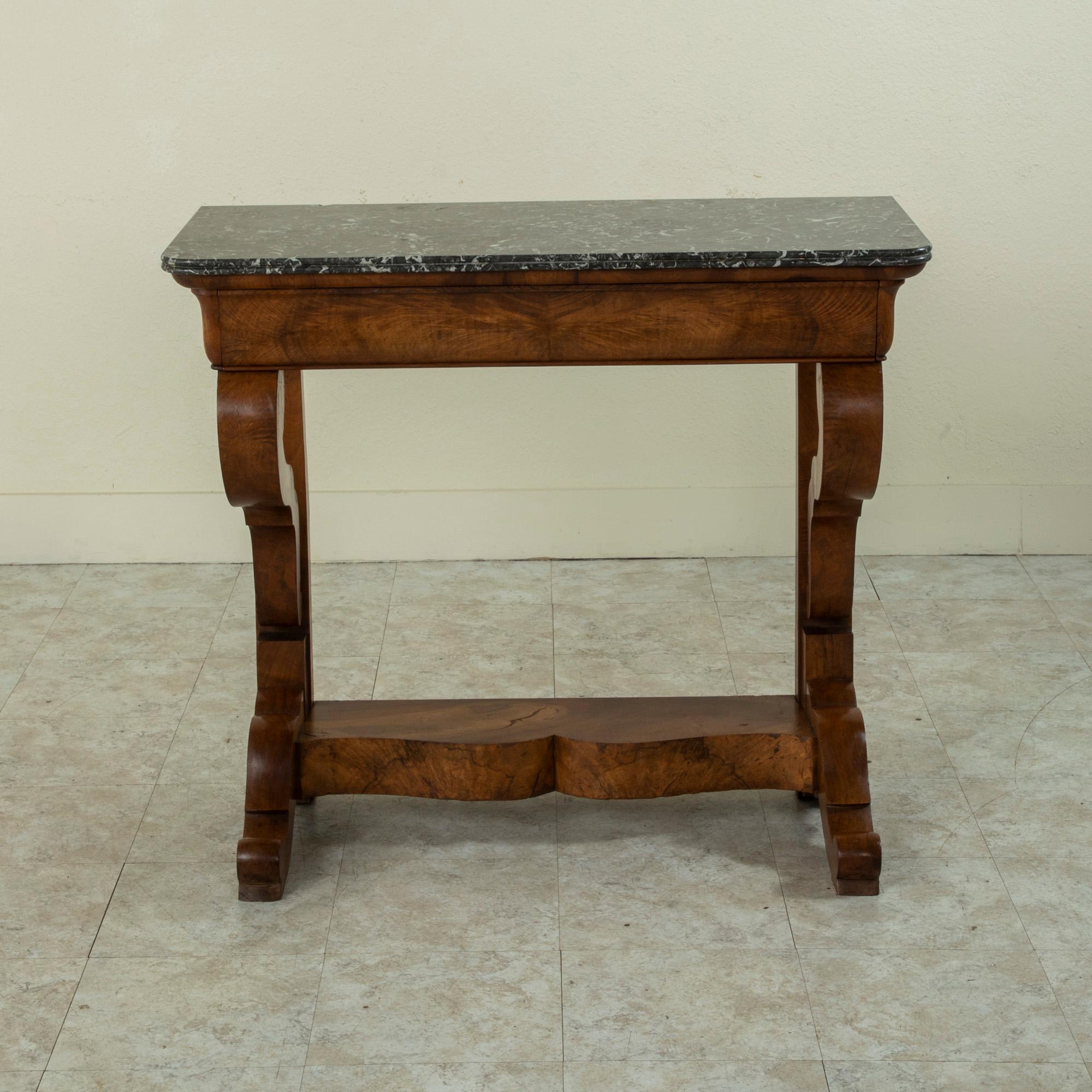 This early nineteenth century French Restauration period console table is constructed of burl walnut and is finished with a beveled Saint Anne marble top. A single drawer seamlessly forms the front of the upper apron. Two curved supports join the