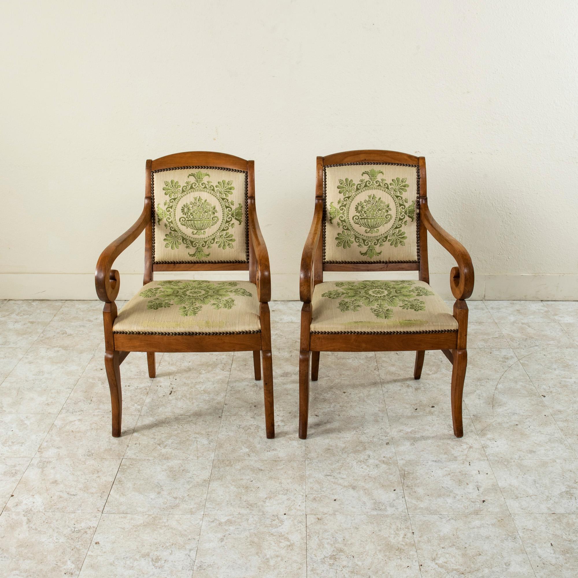 This pair of French Restauration period walnut armchairs from the early nineteenth century features scrolling arms and gently curved legs. The back legs are flared 