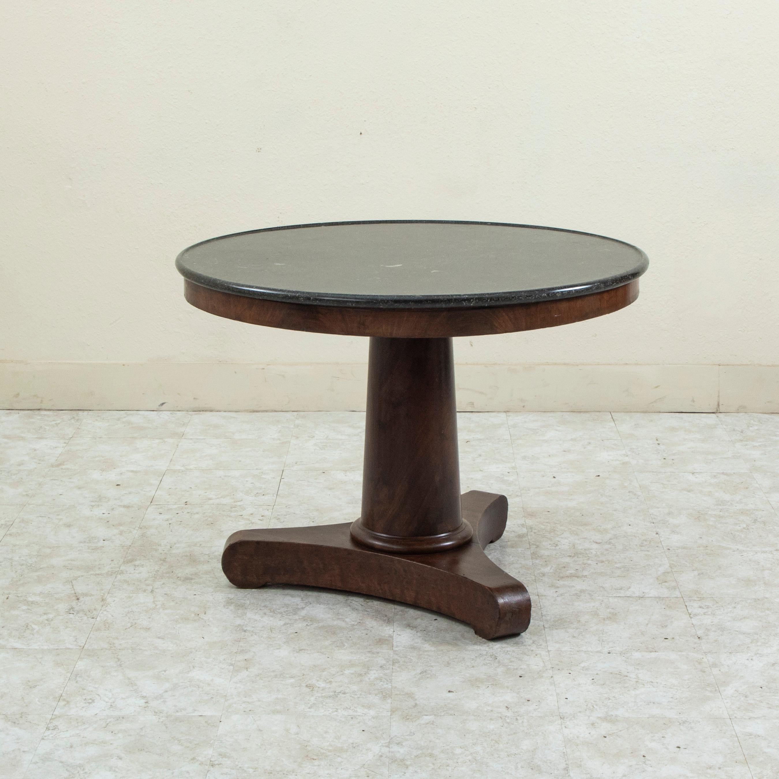 This early nineteenth century French Restauration period mahogany gueridon or pedestal table features a 39 inch diameter beveled top of black marble. The marble displays various fossils as well as white veining., A large central pillar supports the