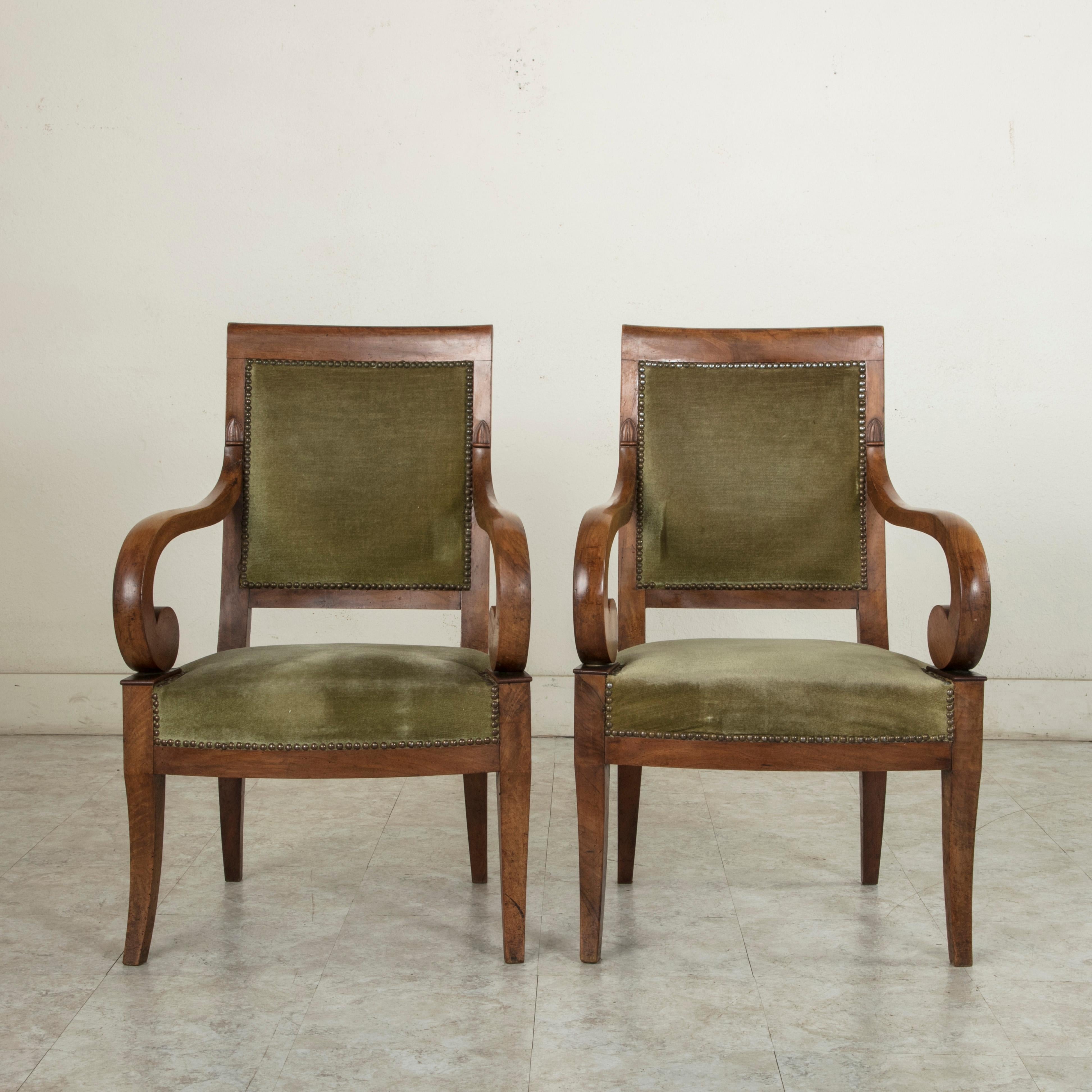 This pair of French Restauration period walnut armchairs from the early 19th century features scrolling arms and gently curved legs. The legs are flared 
