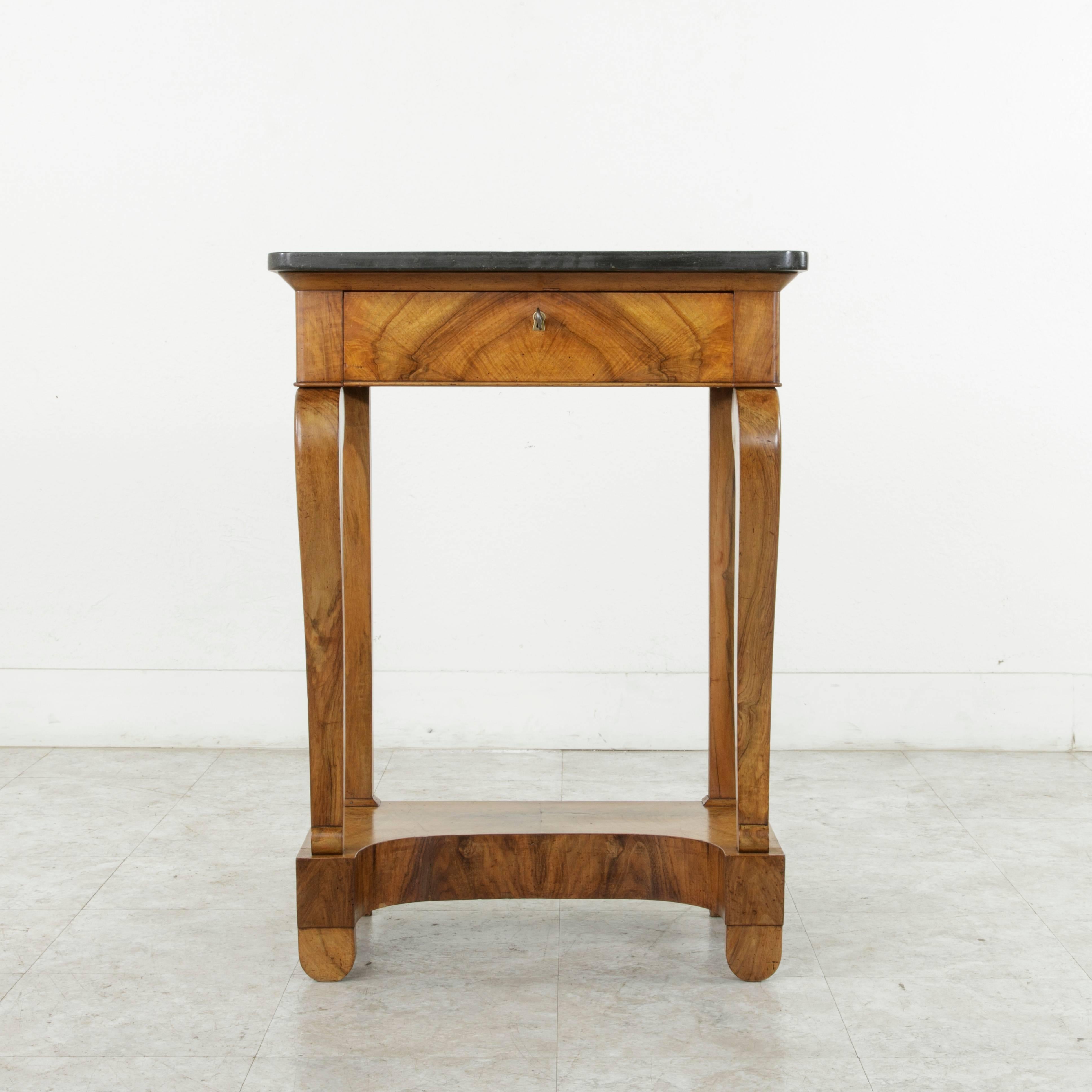 This small-scale early nineteenth century French restauration period console table features stunning bookmatched walnut and is finished with a black marble top. A single drawer of dovetail construction seamlessly forms the front of the upper apron.