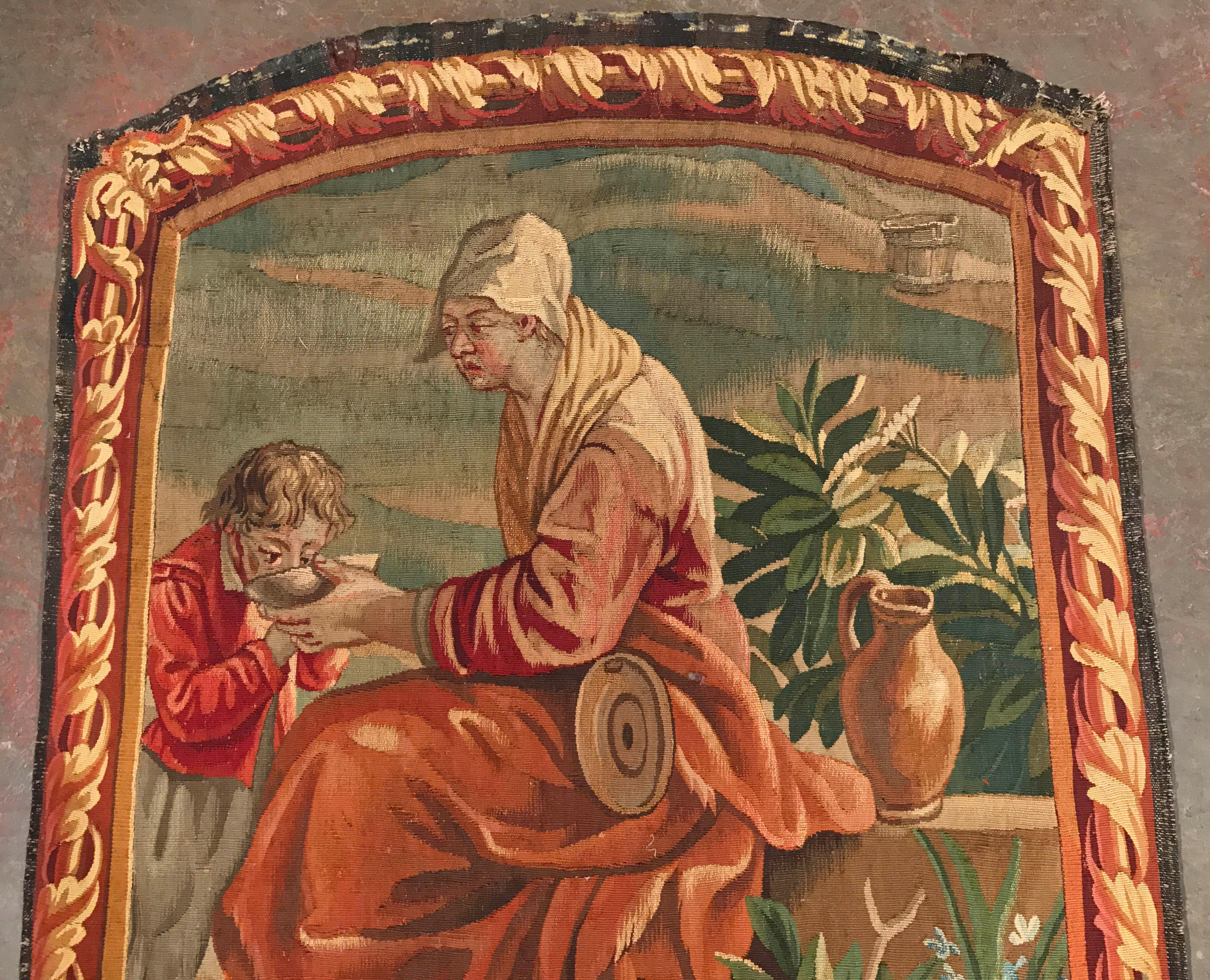 Cette tapisserie ancienne et colorée a été tissée à la main à Aubusson, en France, vers 1820. La tenture murale représente une femme donnant un bol de soupe à un jeune enfant ; l'œuvre d'art est agrémentée d'une bordure tissée en trompe l'œil de