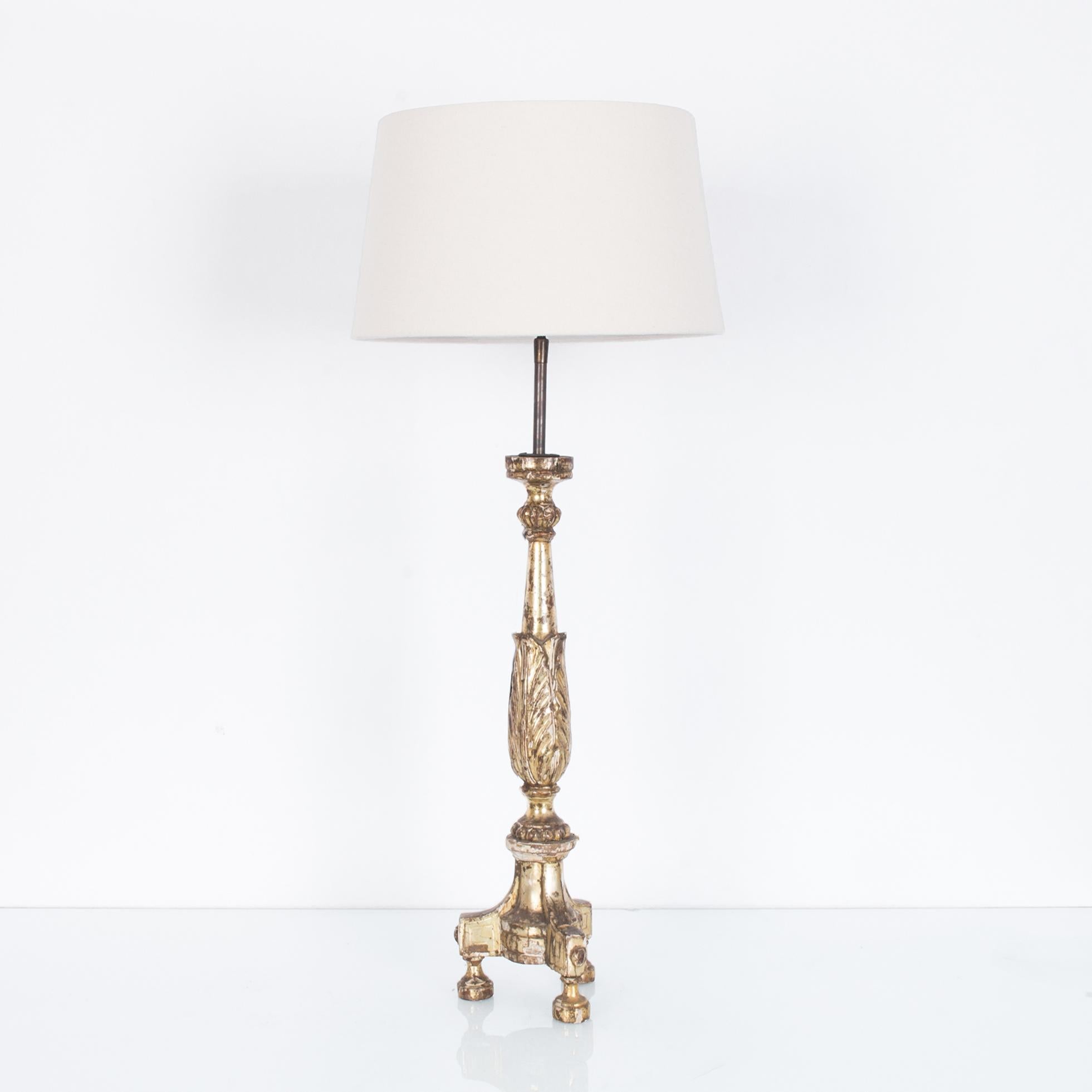 A gilded wooden table lamp from France, circa 1800. A repurposed rococo candlestick supports an empire lampshade in natural white. Three pedestal feet elevate a slender column embellished with an arrangement of carved leaves. The bright gilded