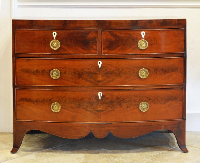 This elegant English bow front chest of drawers features a polished top above two short and two long drawers with ornate brass pulls and recessed bone shield form escutcheons. The chest stands on splayed legs connected by a shaped apron.