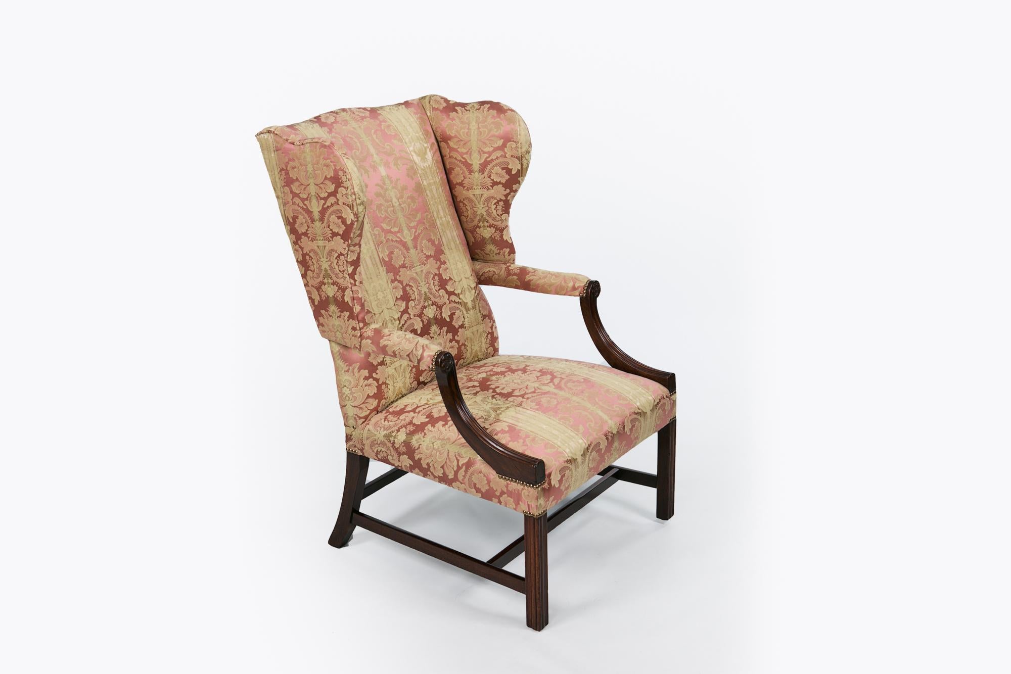 Early 19th century George III mahogany upholstered wing chair, the shaped back with outscrolled wings raised above padded arms with reeded terminals with flower head motif raised over squared moulded leg joined by a H-stretcher.