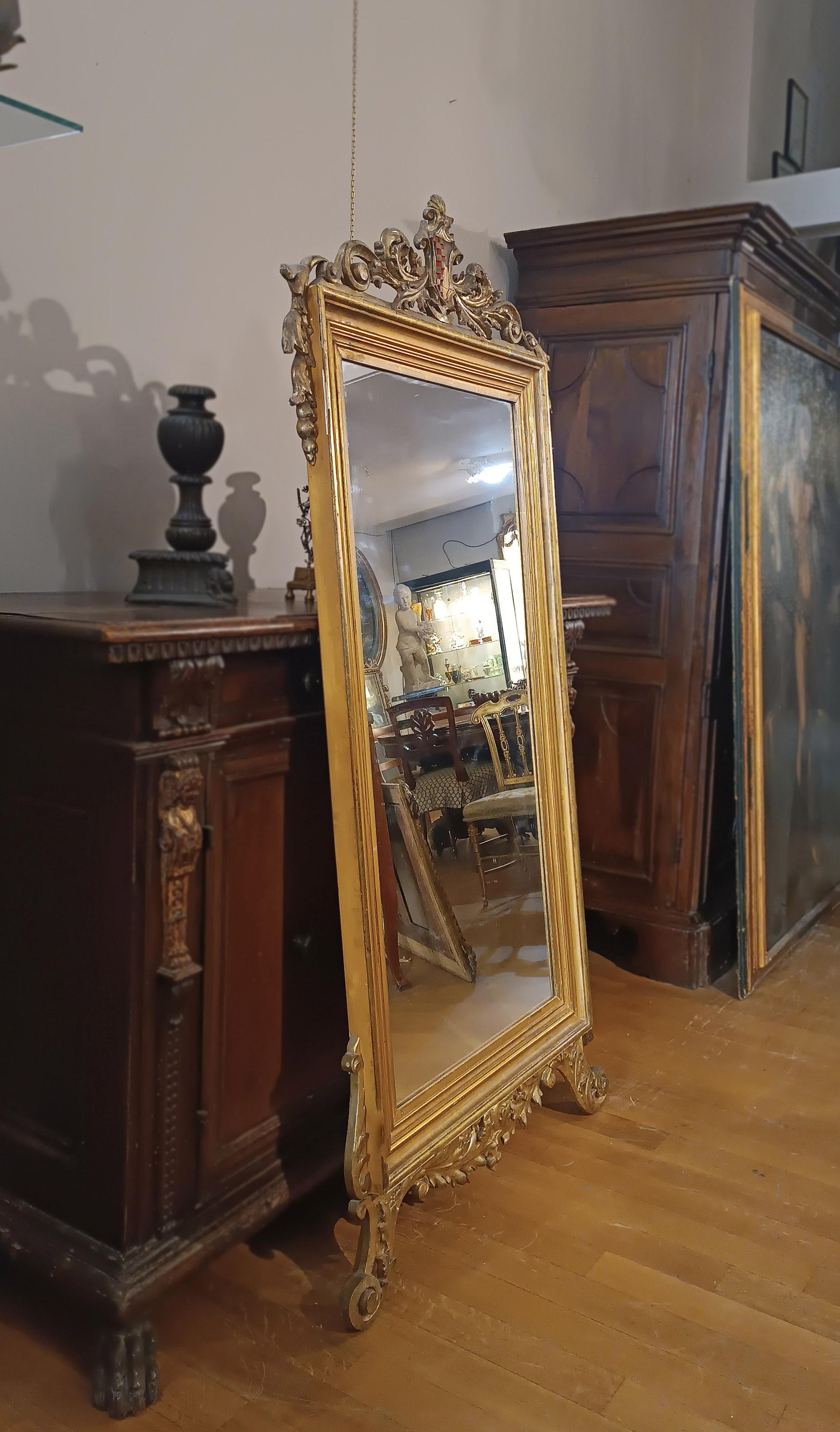 Miroir rectangulaire élégant et raffiné en bois de pin sculpté et doré. Le miroir est soutenu par des pieds feraux décorés d'élégantes spirales végétales, qui ajoutent une touche de raffinement. Sur la partie supérieure, des sculptures ont été