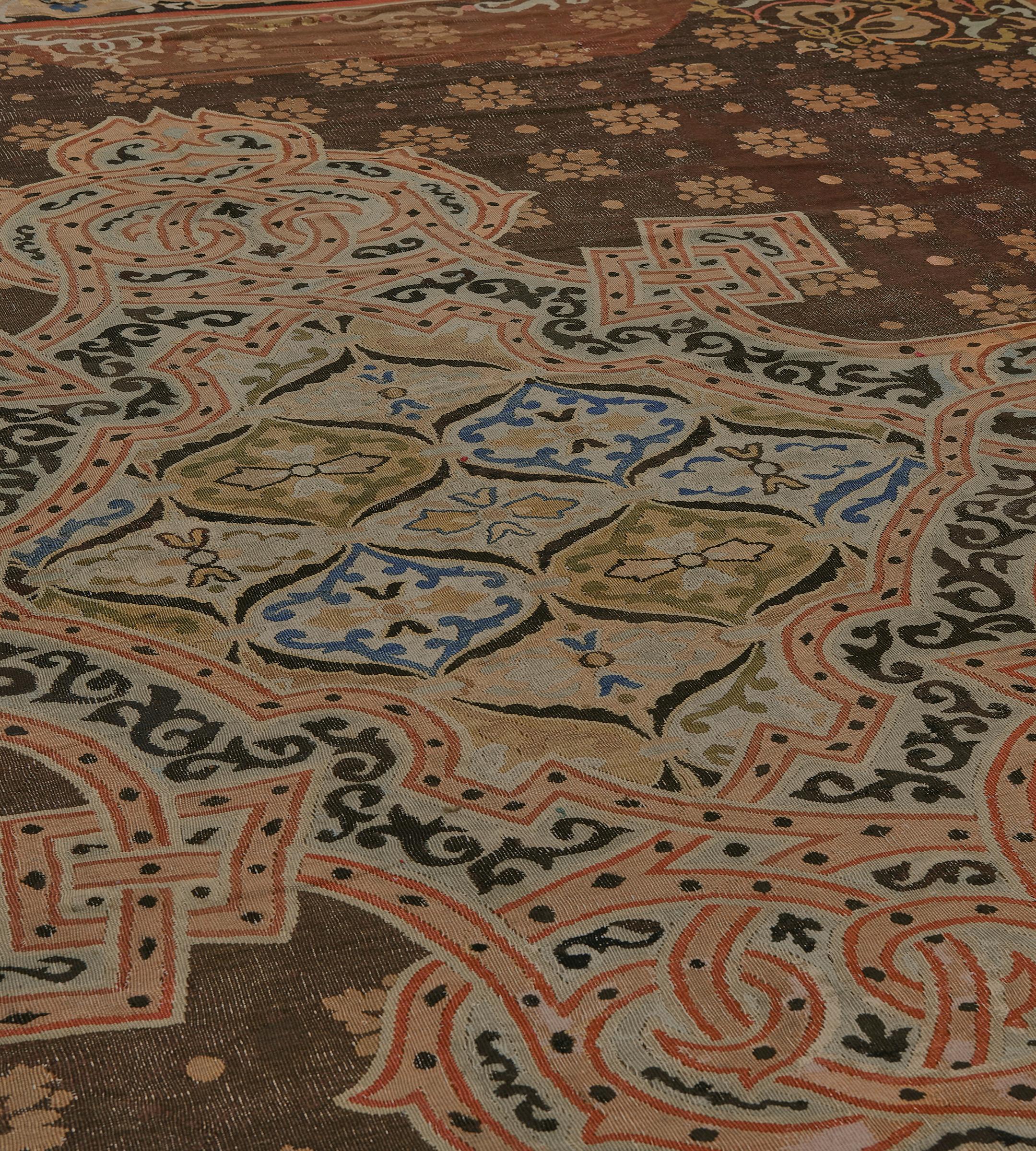 This antique French Aubusson rug features a tobacco-brown field with diagonal rows of light terracotta-orange floral motifs and a delicate similarly coloured scrolling outer band with an onion shaped lozenge in each corner around a large ivory