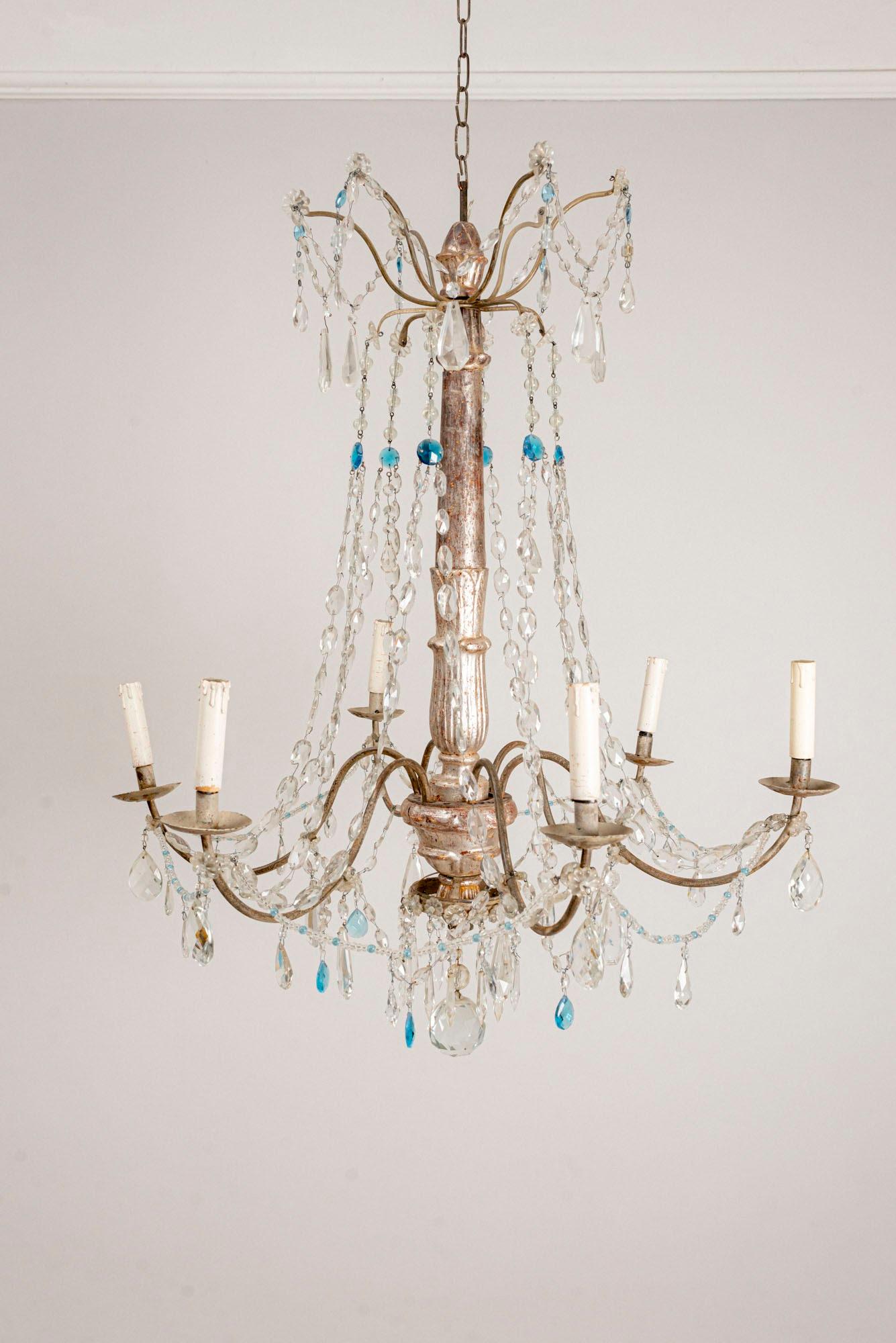 Early circa 19th century Italian chandelier with a silver gilt carved wood central column. 
This charming chandelier has six metal arms with swags of clear and occasional blue glass drops. 
It dates to circa 1820s and is currently unwired but has