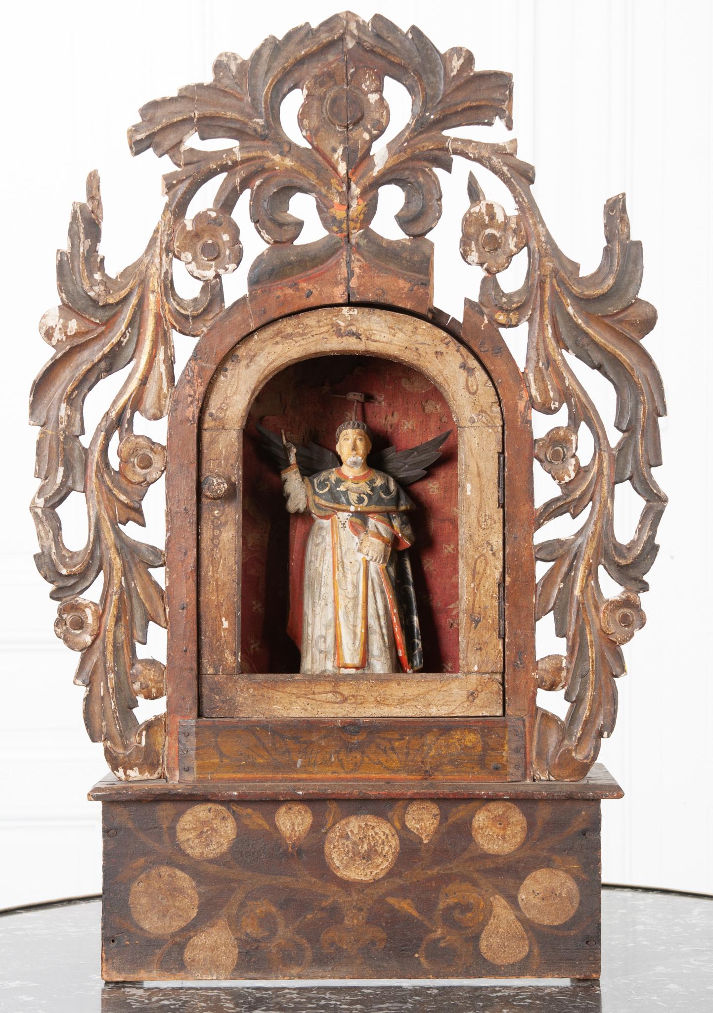 This early 19th century Italian altar is made of worn painted wood and worn fabric details. This is a highly decorative altar for the unknown Catholic saint carved in the center; designated by his upward pointing finger, wings, and attachment where