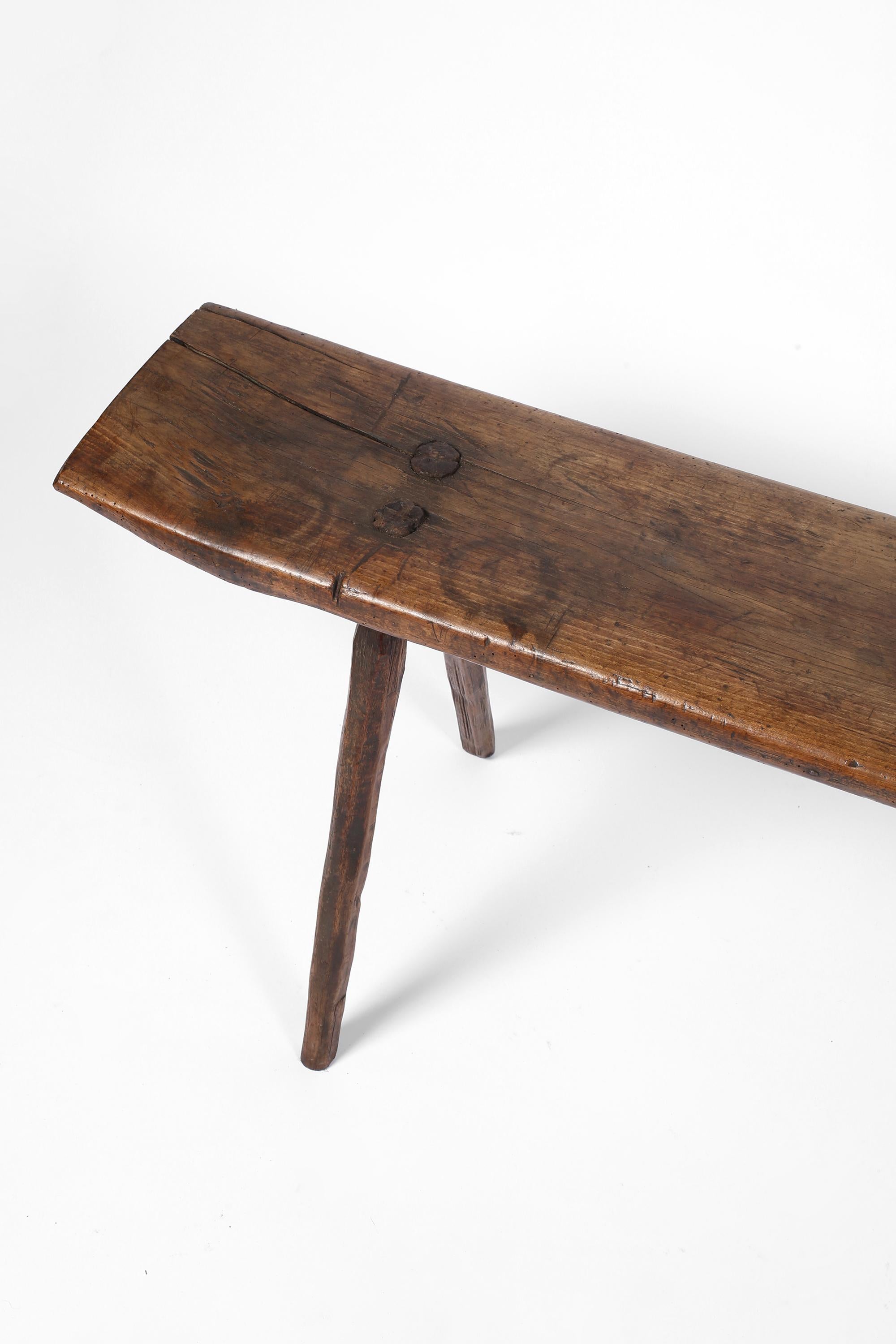 An early 19th century primitive mountain bench constructed from a single plank of richly patinated oak. Retaining its original whittled and attractively splayed legs, with characterful historic iron staple repairs to each end. Italian, c. 1800.