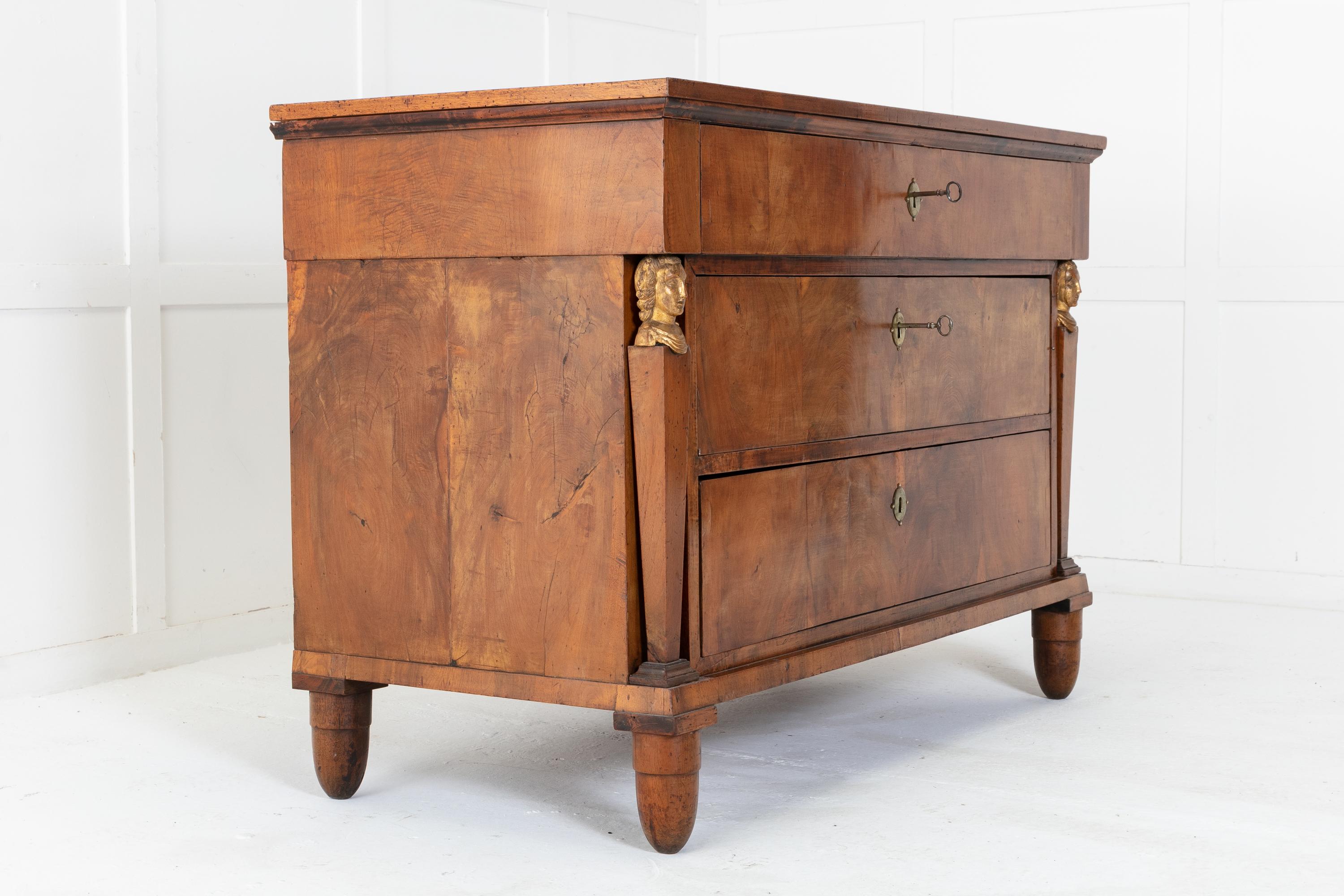 Early 19th century Italian, three drawer, walnut commode. The top sits above a long drawer. A further two graduated drawers below are flanked by square, tapering pillars topped with gilded maiden heads. Each drawer has brass escutcheons with working