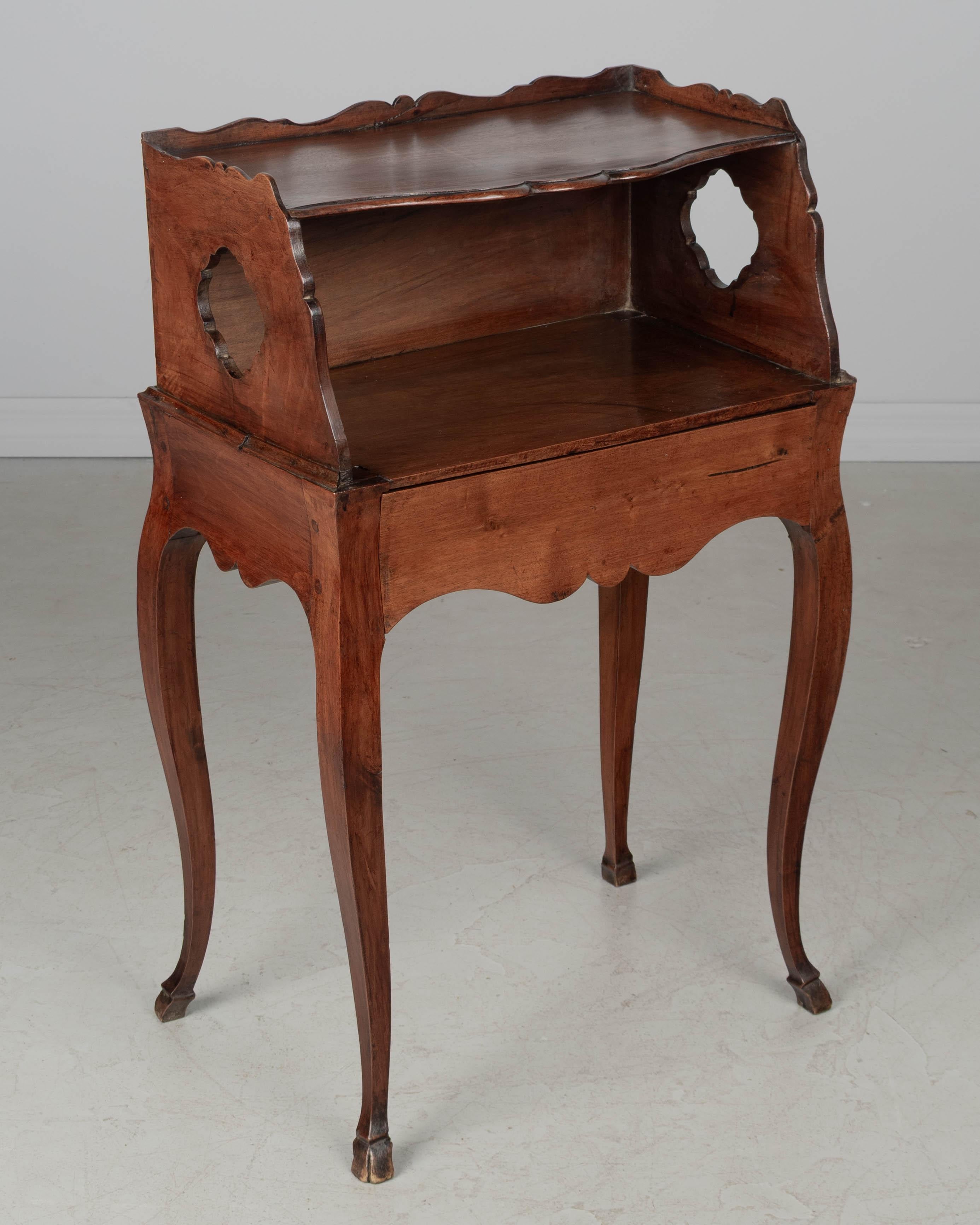 An early 19th century Louis XV country French side table or nightstand made of solid walnut, with pierced quatrefoil shaped cut-outs on the sides. Open niche with dovetailed drawer hidden in the scalloped apron. Beautiful curved form with nice