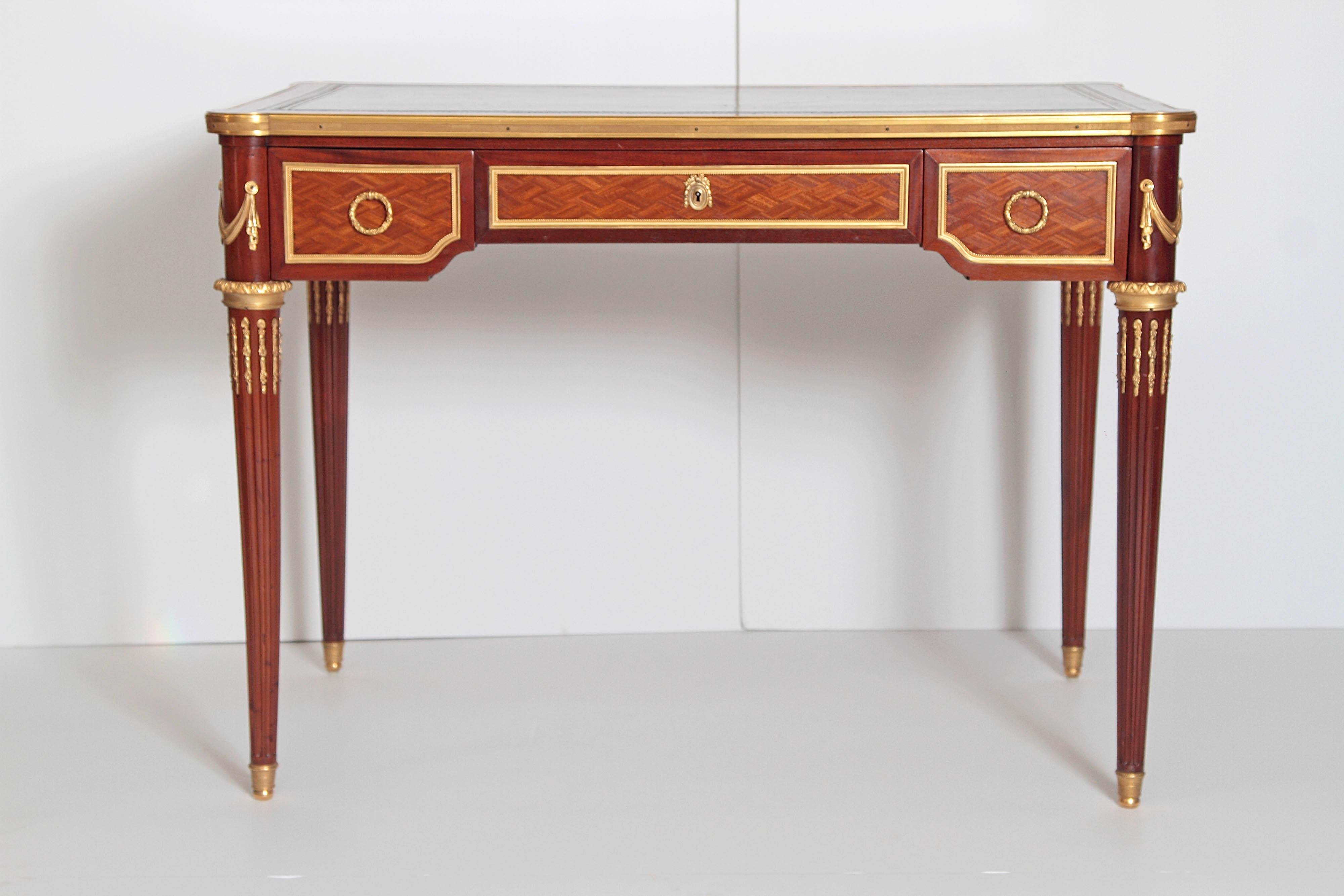 A French Louis XVI style writing table of mahogany. Two small drawers flank larger center drawer. Parquetry inlay on drawer fronts. Green leather top with gilt stamped decoration. Reeded tapered legs with gilt bronze mounts, early 19th century,