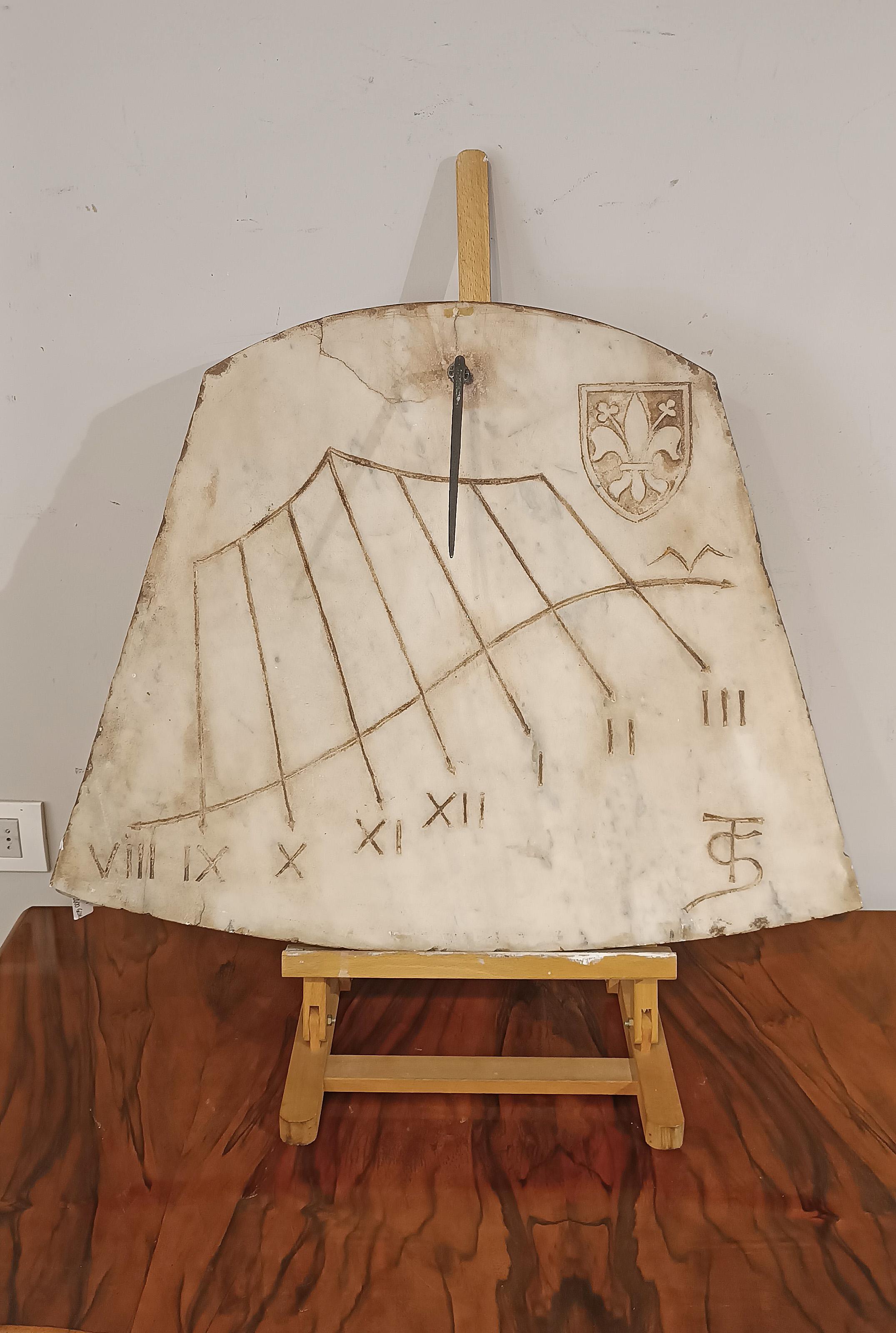 Ancient sundial in carved white marble, from a historic Florentine palace. The sundial features the famous Florentine lily carved at the top right, while the sculptor's monogram is at the bottom right. At the top there is an iron harpoon that marks