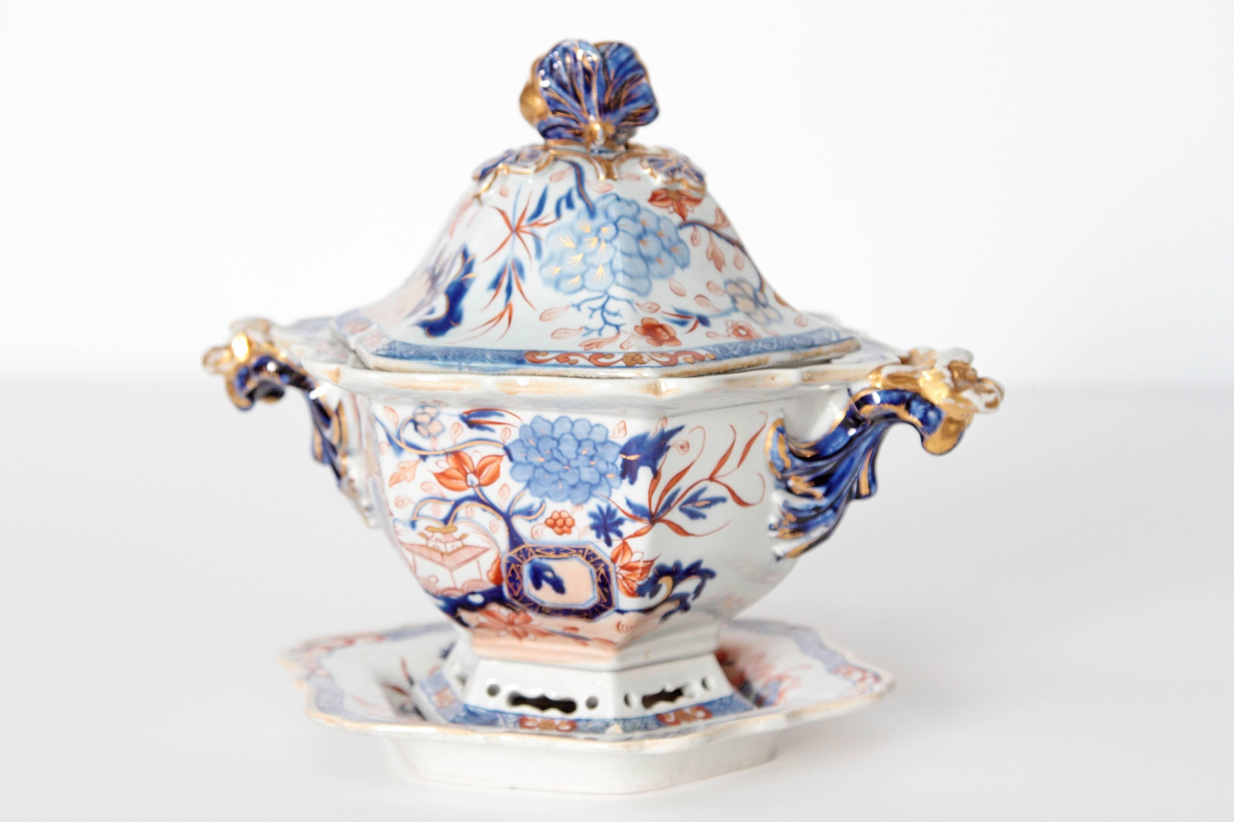 A English soup tureen with under plate decorated with a colorful Imari design. Overall floral scene with pagodas in vibrant blues, orange and gold. Glazing chip to one corner and small missing piece on finial (circled in images), Under plate, tureen