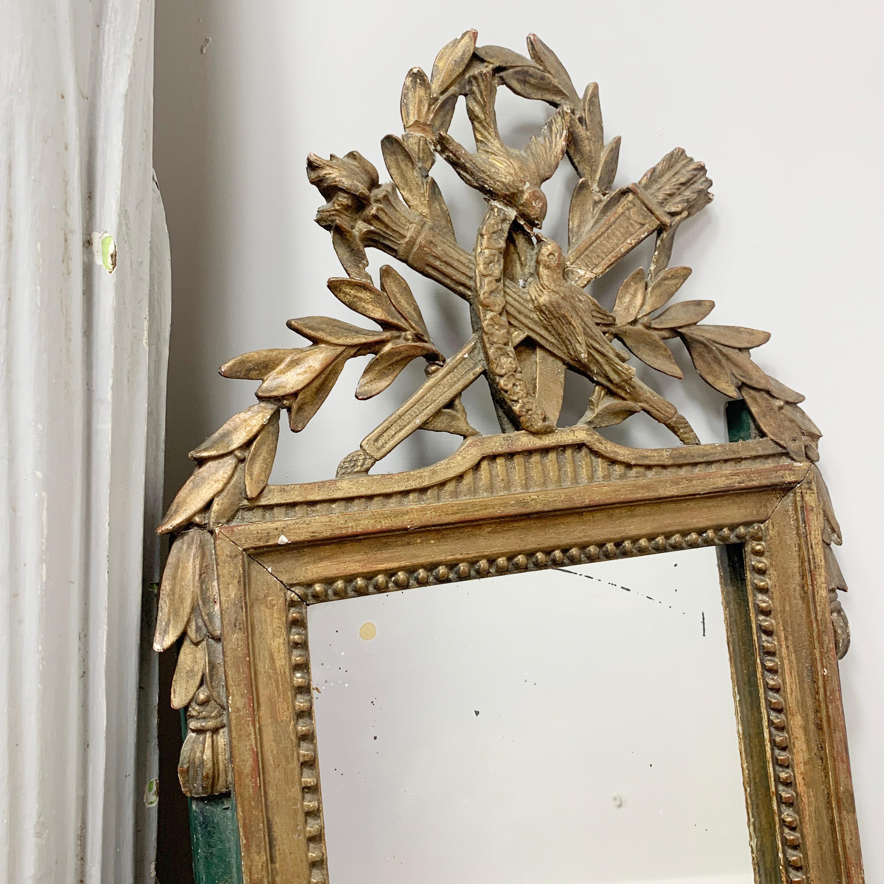 An early 19th century mirror petite, in the Louis XVI style,
Circa 1830
Gilded gesso over carved wood
The frame is gold with bottle green side details, it is adorned with a crest of carved wreath, birds and quiver
Patina and wear commensurate