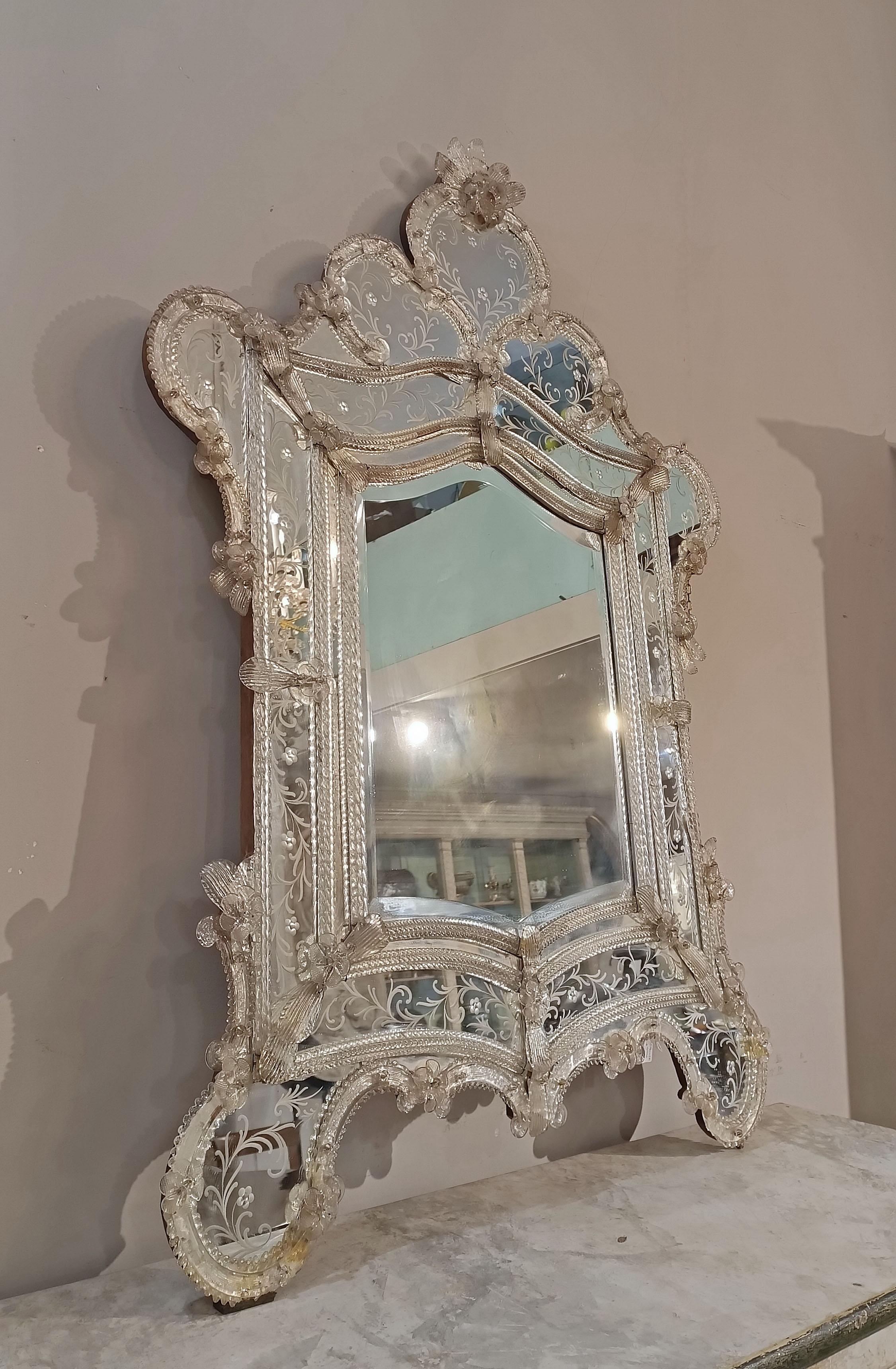 Elegante y refinado espejo de cristal incoloro de Murano. El marco está bellamente decorado con flores, cuentas y sinuosos giros de cristal, que rodean espejos grabados con motivos florales, lo que añade gran elegancia a la pieza. La cresta, también