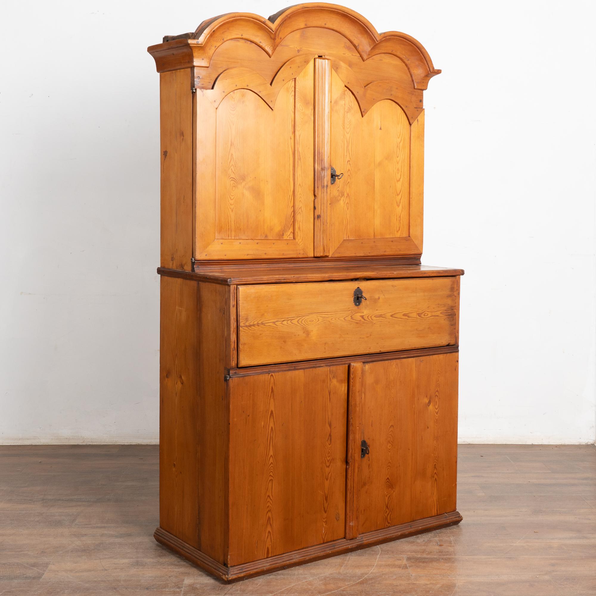 Este armario de pino es un magnífico ejemplo de la artesanía rústica noruega, que incluye el tradicional bonete curvo o corona y puertas con paneles. 
Fíjate en la sección central abatible, que permite utilizarla como espacio de almacenamiento