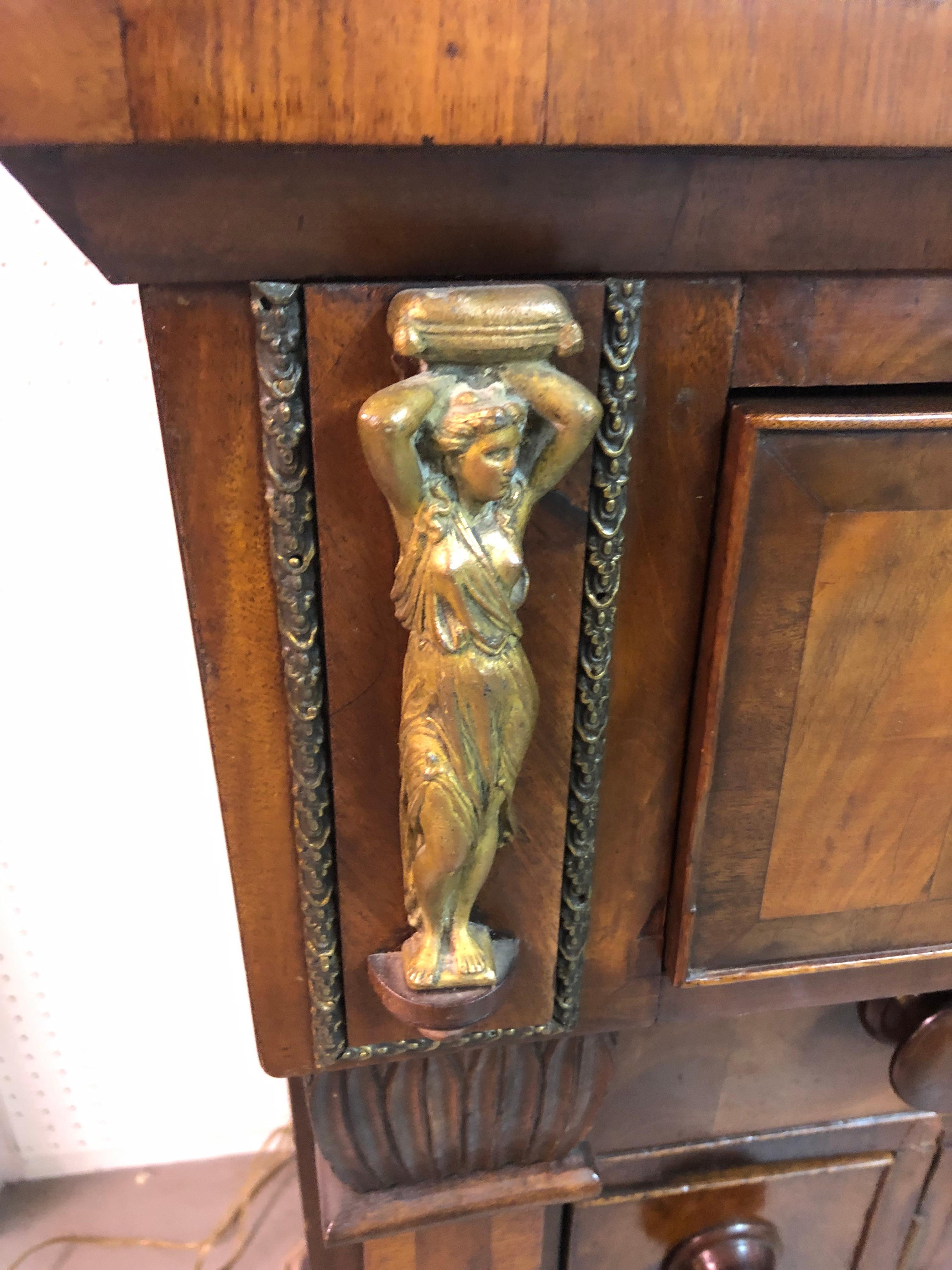 An old world mahogany Empire sideboard having molded top, cockbeaded drawer fronts, and gorgeous brass neoclassical figural mounts. There are two paneled doors with storage inside, two liquor bottle storage compartments, two drawers with one lined