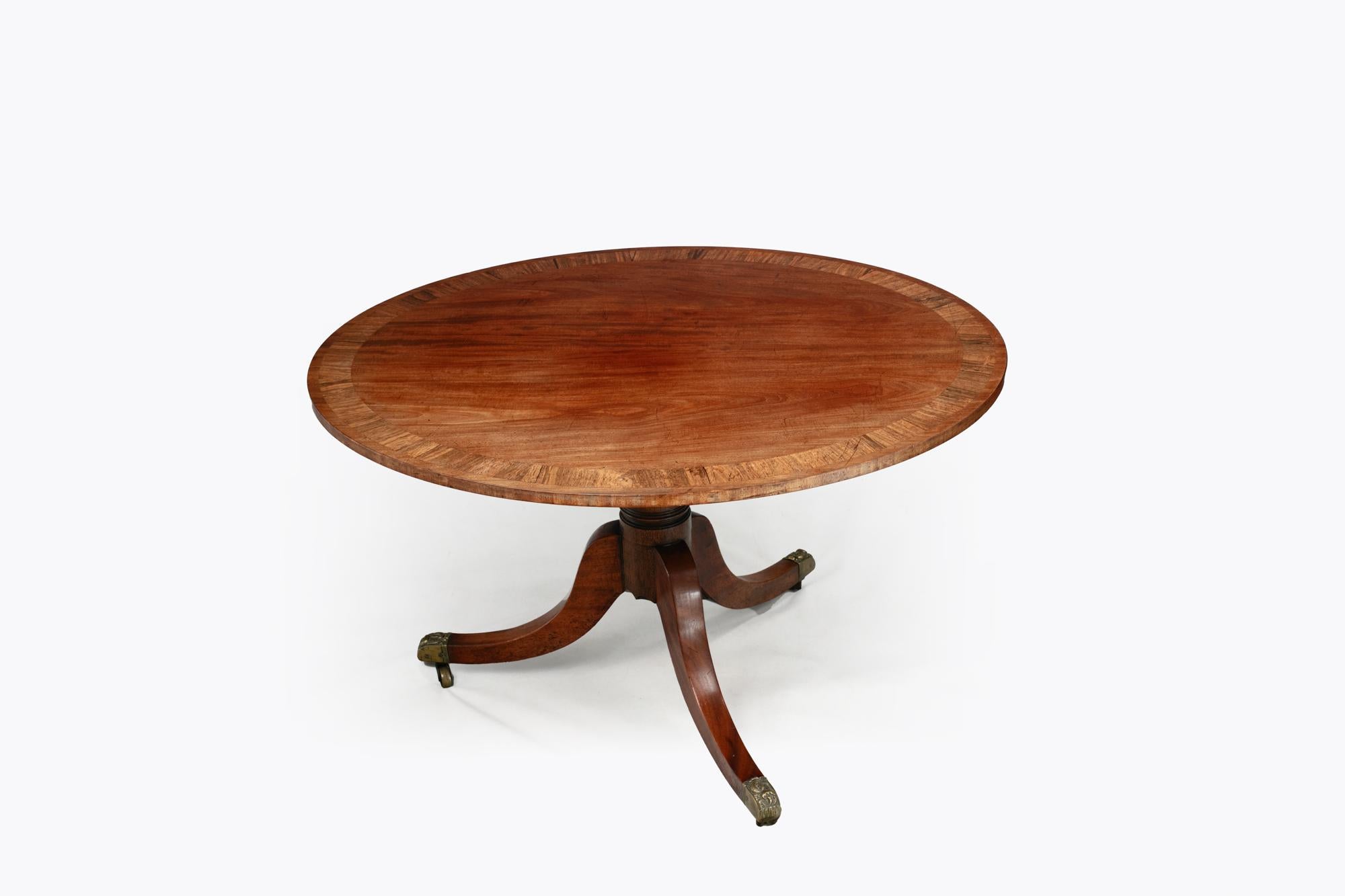 Table à déjeuner ovale du début du 19e siècle en acajou avec bandes transversales en bois de rose, reposant sur un pied supporté par trois jambes évasées sur des roulettes en laiton. Datant d'environ 1820